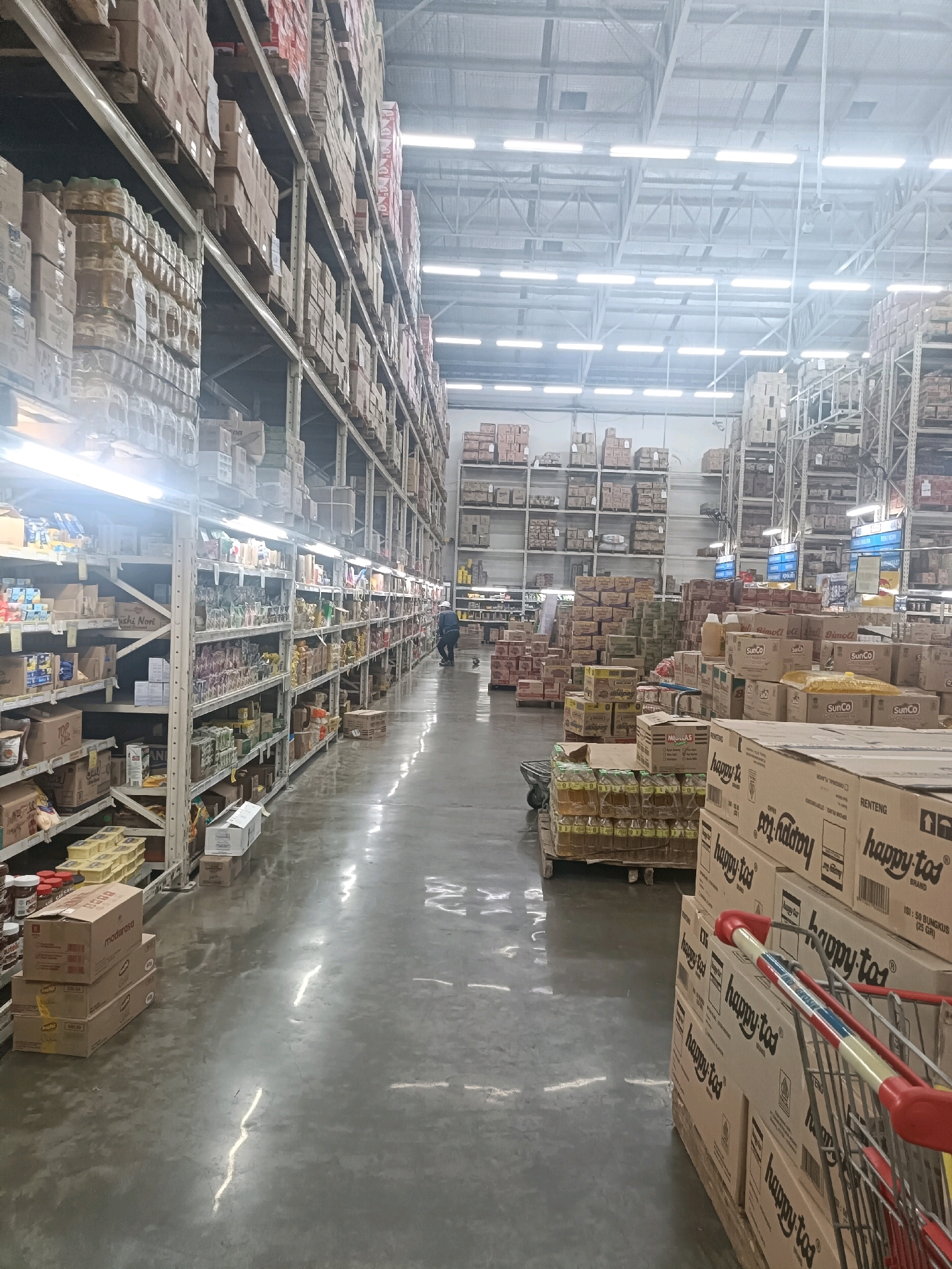 The image depicts a wide aisle within a large warehouse-style retail store, likely a hypermarket or wholesale club, in Semarang, Indonesia. Towering shelves on both sides of the aisle are filled with numerous cardboard boxes and packaged goods, stretching deep into the distance. The shelves are organized and stacked with items ranging from food products to household goods, all densely packed. The lighting is bright and uniform, emanating from long fluorescent fixtures on the high ceiling, which is supported by a metal framework. The floor is a polished concrete surface, reflecting the overhead lights and creating a glossy appearance.
In the mid-ground, a person dressed in dark clothing and wearing a backpack is walking away from the camera down the aisle. Further down the aisle, another person in dark clothing with a lighter colored backpack is visible. In the foreground, to the right, a shopping cart with a red handle is positioned alongside stacks of boxes labeled "happy tos." To the left, several pallets of bottled goods are stacked on the floor, some wrapped in plastic. Scattered boxes and merchandise are also visible on the floor along the shelves. The overall impression is one of a well-stocked, large-scale retail environment designed for mass distribution.