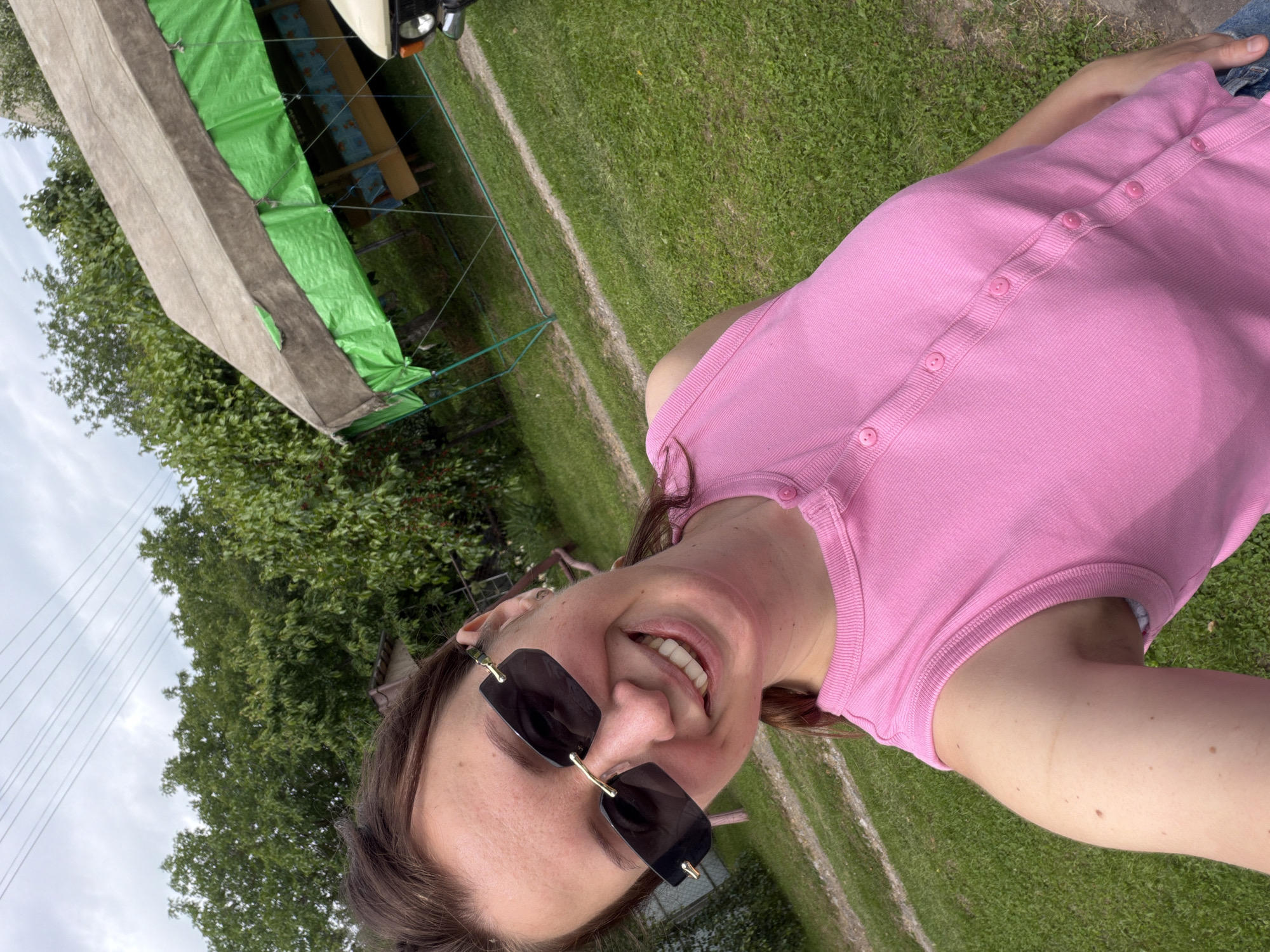 A medium shot captures a young woman in sunglasses and a pink sleeveless top, smiling widely and looking at the camera. She is outdoors, with a grassy area and trees in the background. A tent-like structure, possibly a gazebo, with green fabric sides is visible to her right. The scene appears to be in a rural or suburban setting, consistent with a location in Kivshovata, Ukraine. The lighting suggests it is daytime.
