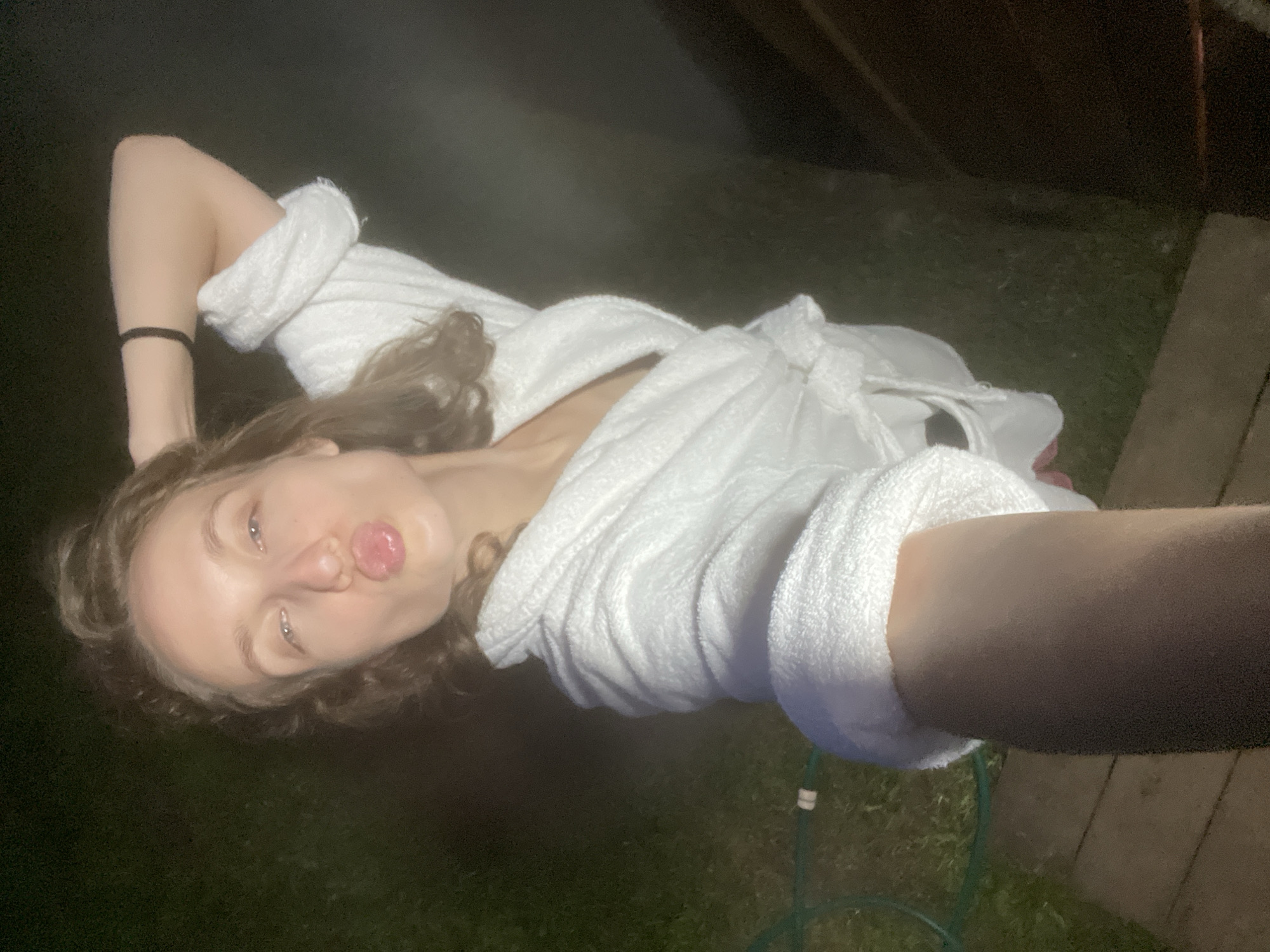 A person, with light skin, is taking a selfie at night, possibly in Sharkan, Russia. They are wearing a white terrycloth robe and have their hair pulled up. Their right arm is bent behind their head, and their left arm is holding the phone, out of frame. They are puckering their lips, as if for a kiss. The background appears to be outdoors, with grass visible and possibly a wooden deck. The lighting is dim, suggesting it is nighttime. There are no other people or animals in the frame. The overall scene is casual and intimate.