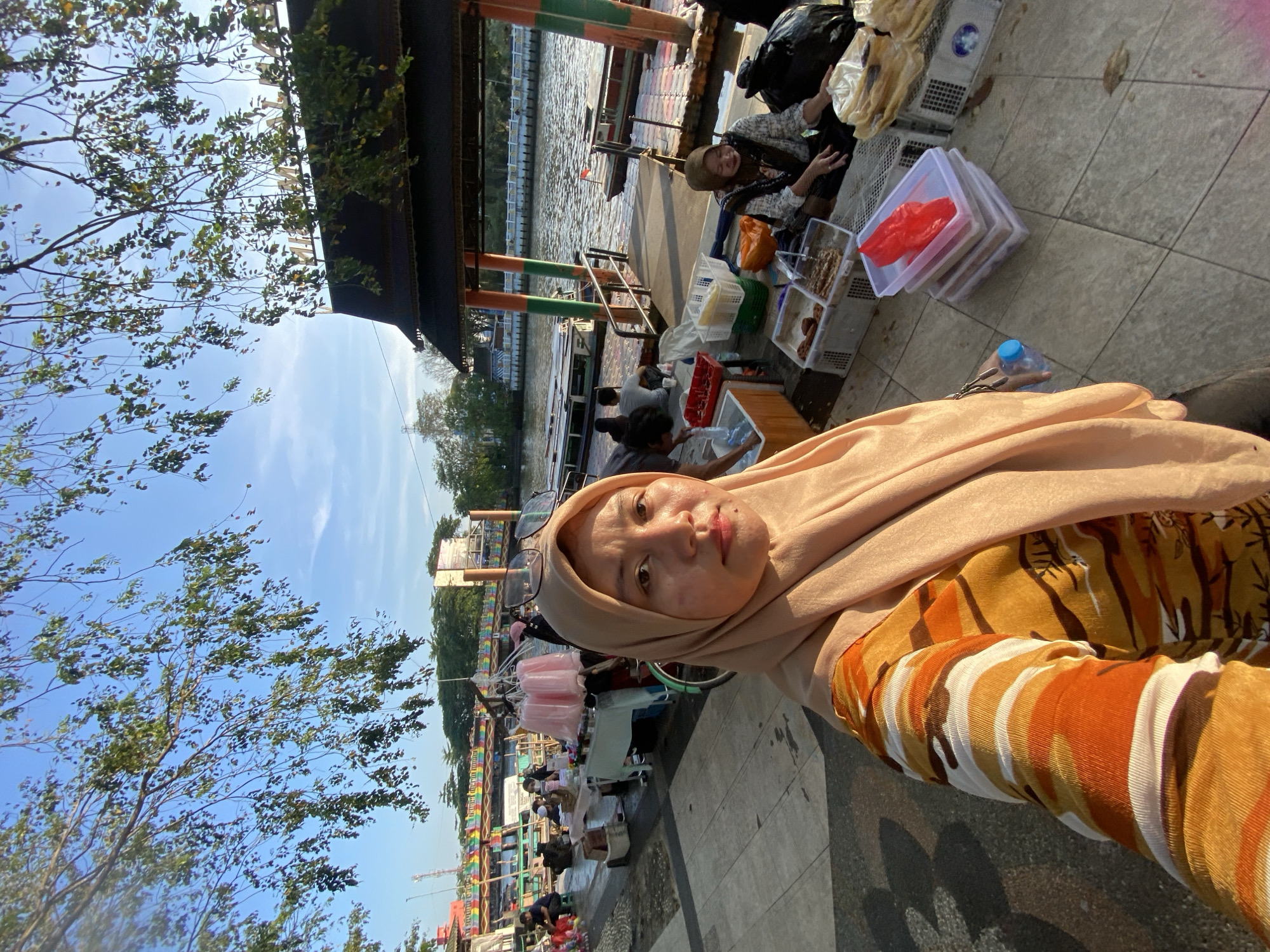 The image depicts a scene in Banjarmasin, Indonesia, likely at a riverside market or promenade. In the foreground, a woman wearing a peach hijab and an orange and yellow striped shirt is taking a selfie. Her expression is neutral, and she is looking directly at the camera. Behind her, the scene unfolds along a tiled walkway next to a river.
Several market stalls are set up along the walkway. One stall features pink cotton candy. Further down, there are various food items displayed in clear containers and baskets. Some people are seated or standing around these stalls, appearing to be vendors or customers.
The river is visible in the middle ground, with several boats docked along a floating platform. The water is calm, reflecting the sky and the surrounding structures. On the far side of the river, there are buildings and trees, suggesting an urban environment.
The overall atmosphere appears to be that of a bustling outdoor market on a sunny day, with the river serving as a central element of the location. The architecture of the covered stalls, with their dark roofs and ornate trim, hints at traditional Indonesian design. The presence of boats indicates that river transportation is common in this area. The clear blue sky and dappled sunlight suggest it is morning or late afternoon.