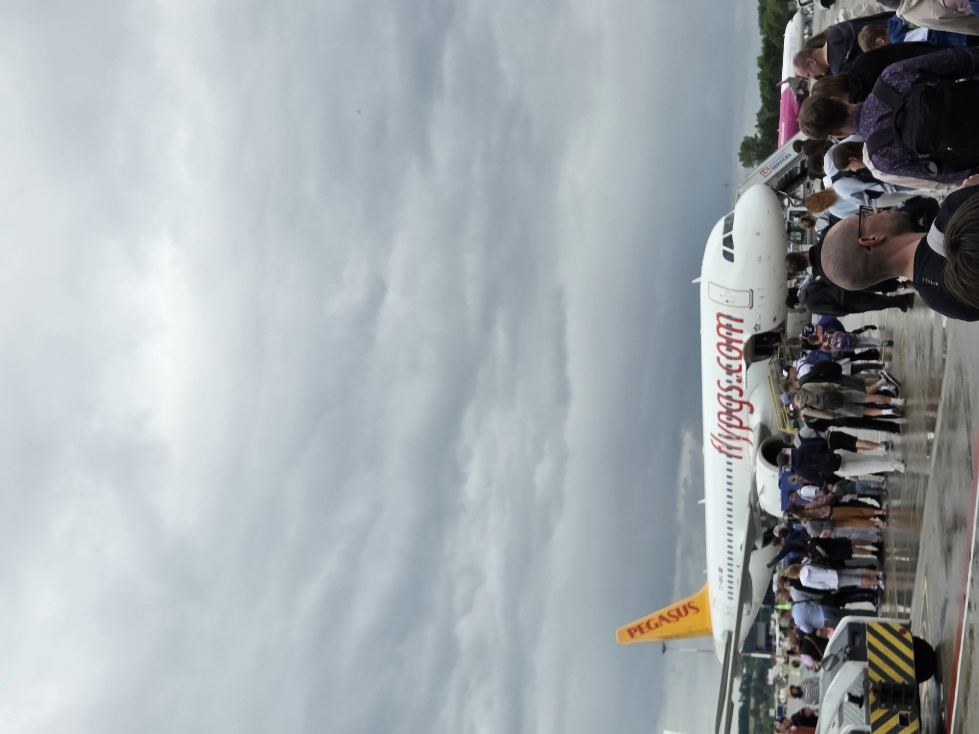 The image depicts a scene at an airport, specifically on the tarmac near a Pegasus Airlines airplane. The sky is overcast with dark clouds, suggesting a possibly rainy day, and the tarmac appears wet, reflecting the overcast sky. A line of passengers, dressed in casual attire, are gathered near the airplane's stairs, seemingly waiting to board or having just disembarked. The Pegasus airplane, with its distinctive yellow tail fin and "flypgs.com" branding, is prominent in the foreground. In the background, another airplane, possibly of Wizz Air given its pink livery, is visible. Airport ground equipment, including a yellow and black striped vehicle, is also present on the tarmac. The overall impression is one of an active, though potentially damp, airport operational moment. The location is identified as Balice, Poland, which is home to John Paul II International Airport Kraków–Balice.