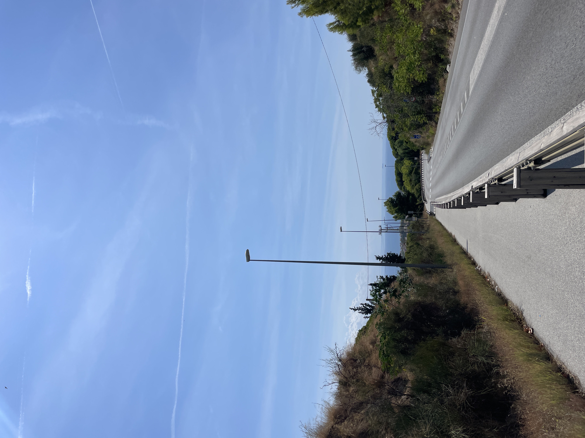 The image depicts a road in Badalona, Spain, on a clear day with a bright blue sky and scattered wispy clouds. The road slopes gently upwards and curves to the right, bordered by a sidewalk on the left and a wooden guardrail on the right. The landscape consists of dry, grassy hillsides with scattered bushes and trees, transitioning to more verdant foliage and larger trees further down the road. In the distance, a body of water, likely the Mediterranean Sea, is visible. Several streetlights are positioned along the road. There are no people or vehicles visible in the scene.