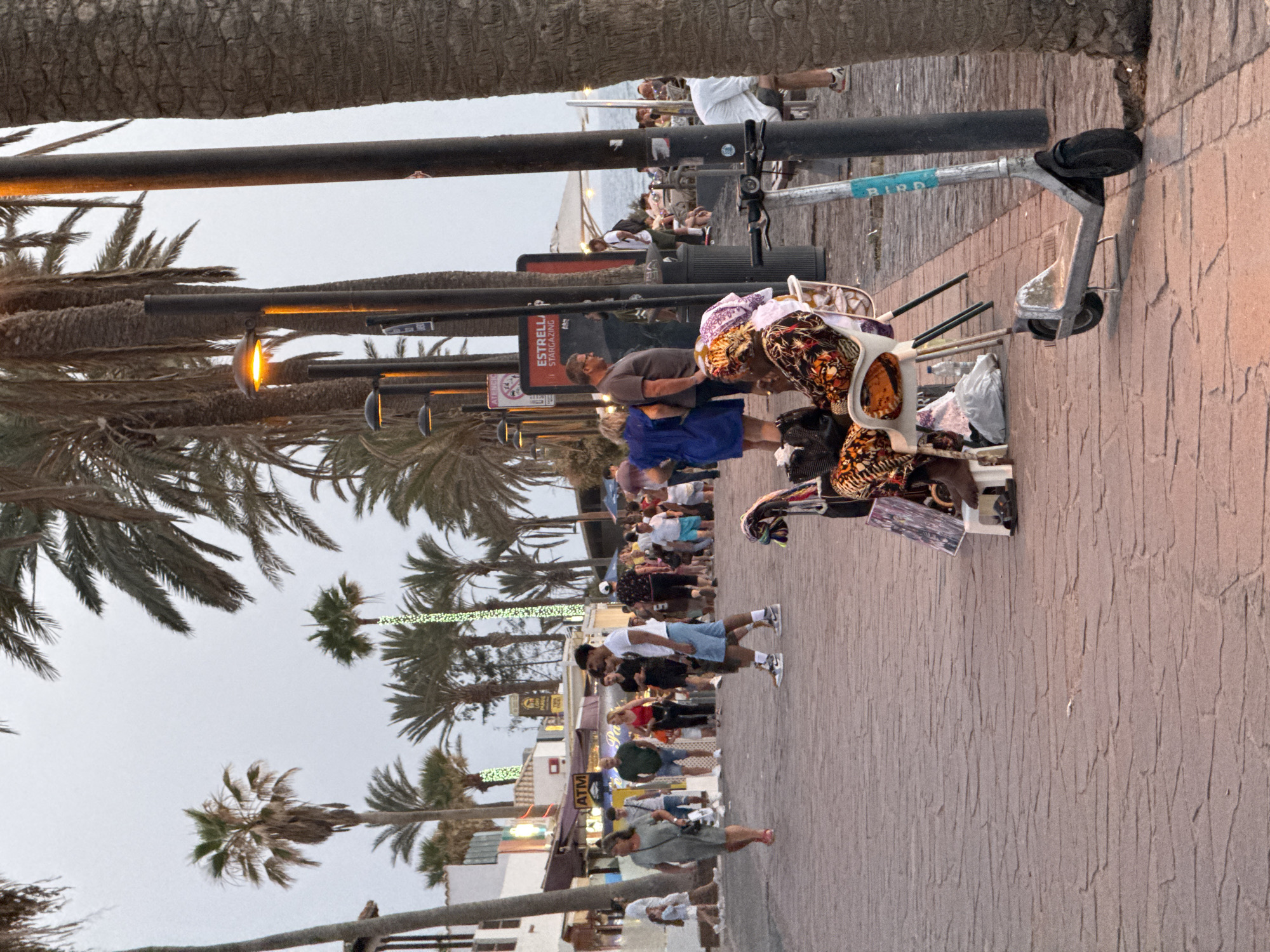 A street scene unfolds on a paved walkway lined with palm trees. The walkway is made of interlocking bricks. To the left, buildings with storefronts, including an ATM, are visible. People are walking along the path in both directions. In the foreground, a woman in a colorful outfit is seated on a white chair, with shopping bags around her. An electric scooter stands nearby. The setting appears to be a coastal promenade in Arona, Spain, suggested by the palm trees and the general atmosphere. The sky is overcast, and the lighting suggests it is late afternoon or early evening.
