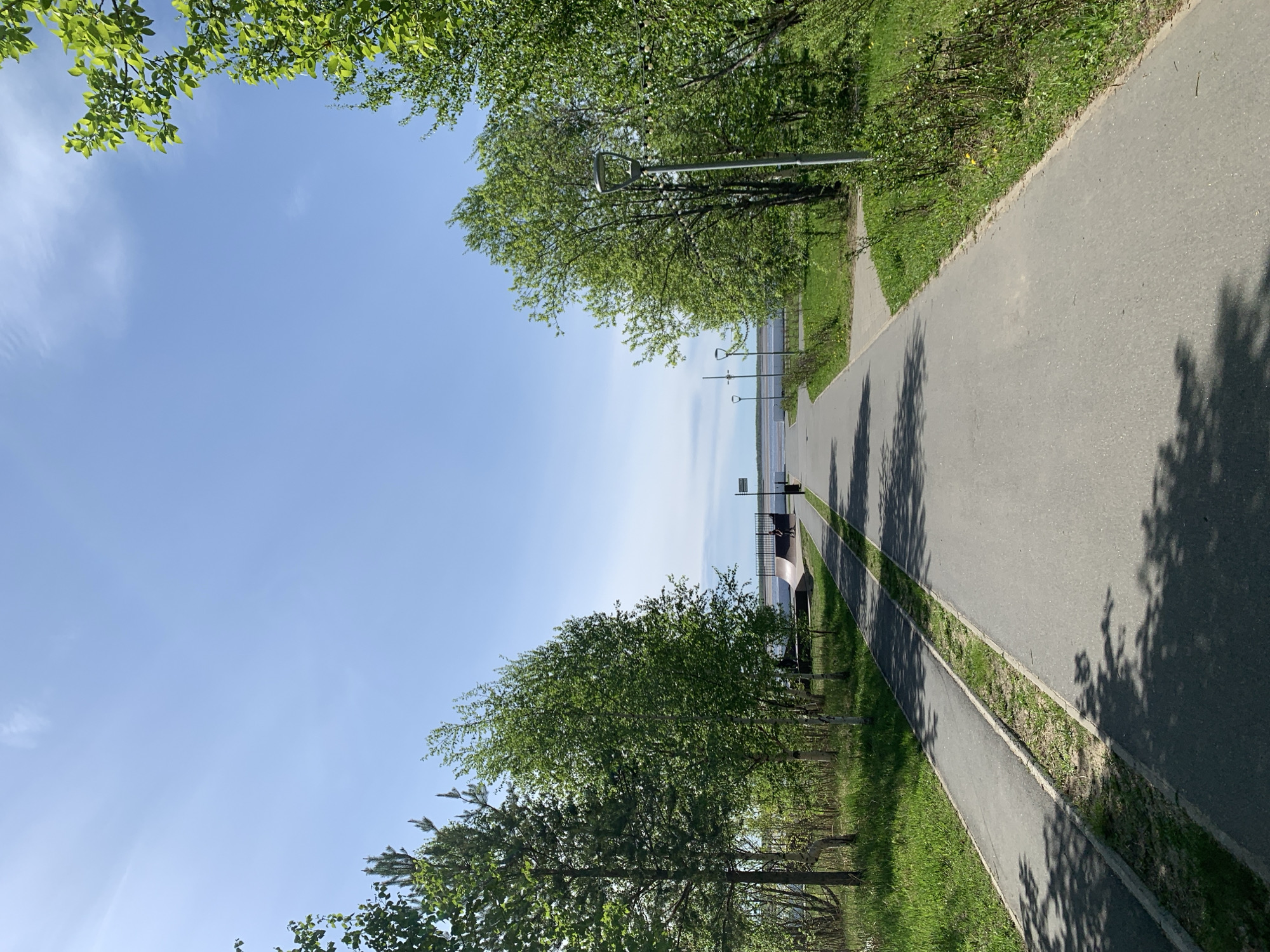 The image shows a paved path in Severodvinsk, Russia, bordered by trees and grass on both sides. In the distance, a body of water is visible, with lampposts along the shore. A small structure with seating is present along the path. The sky is blue with a few clouds. The orientation of the image is vertical.