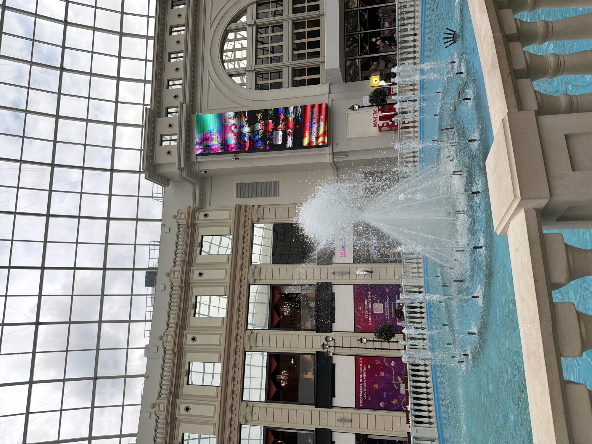 The image captures an interior scene of a large building, possibly a shopping mall or entertainment complex, with a prominent fountain in the foreground. The fountain features multiple water jets spraying upwards into the air, creating a dynamic visual effect against the backdrop of the building's architecture. The water in the fountain is a striking turquoise color.
The building itself appears to have a classical or ornate design, with decorative moldings, columns, and a large glass ceiling that allows natural light to filter in. The ceiling is made up of numerous square panes, suggesting a grand and open space. Below the ceiling, the building's facade displays several levels of windows and illuminated advertisements.
One prominent advertisement is a large, colorful LED screen displaying abstract, vibrant graphics, possibly an art installation or digital advertisement. Other large posters or banners with text are visible on the walls of the building, some featuring images and Russian text. One banner reads "ТВОЙ БИЛЕТ В МИР ПРИВИЛЕГИЙ" which translates to "YOUR TICKET TO THE WORLD OF PRIVILEGES." Another banner says "ПРОГРАММА ЛОЯЛЬНОСТИ «ОСТРОВА МЕЧТЫ»" which means "LOYALTY PROGRAM 'ISLAND OF DREAMS'."
The overall lighting suggests it is daytime, with the natural light from the glass ceiling illuminating the scene. The weather outside, visible through the ceiling, appears overcast. Reflections of the windows and the sky are visible on the glass panes of the building. There are no distinct people clearly visible in the image, though some figures might be present in the reflections of the windows or on the distant advertisements, but they are not a clear subject. The atmosphere appears to be one of a public, well-maintained space, likely designed for leisure or commerce.