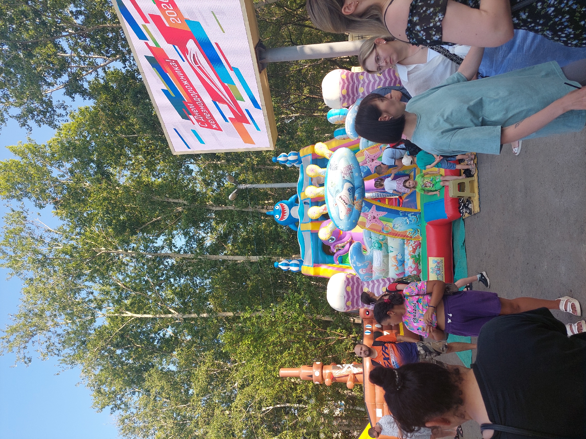 The image depicts an outdoor scene in Komsomolsk-on-Amur, Russia, with a large, inflatable playground structure as the central focus. The playground is brightly colored and features various cartoon characters, including a shark at the top. Several children and adults are present, some of whom appear to be enjoying the playground, while others are observing or walking around. In the background, tall trees form a dense canopy against a clear blue sky. To the right of the playground, a large digital billboard displays a festive message and imagery, likely related to a public event or holiday. The ground is paved, and the overall atmosphere suggests a community gathering or celebration.