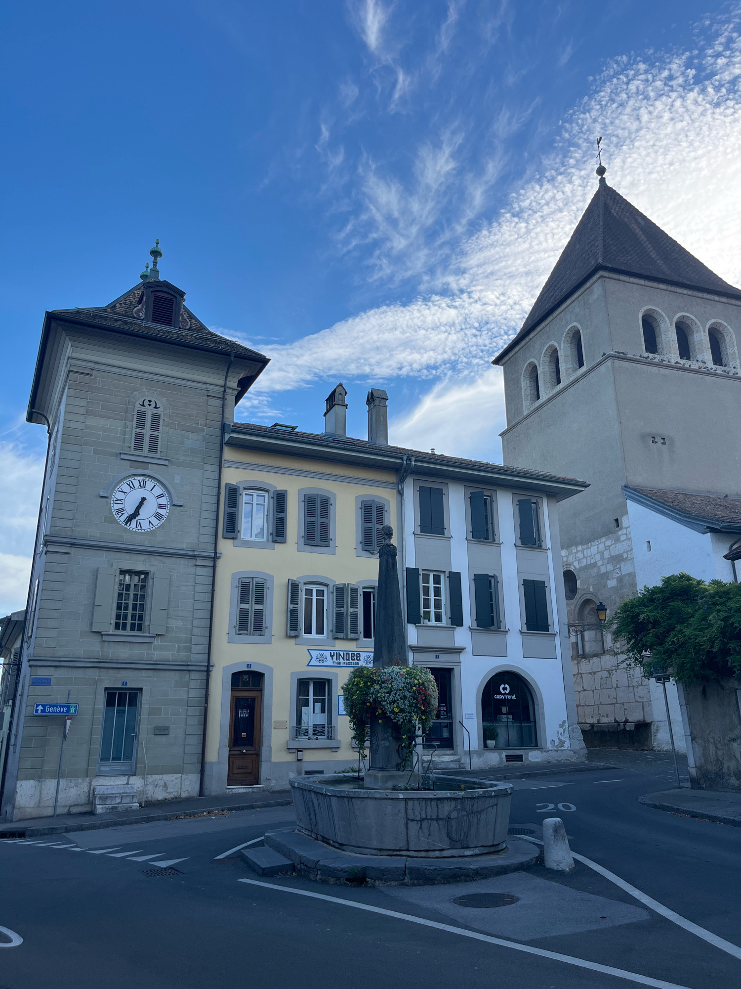 This is an eye-level, medium shot of the Place du Château in Nyon, Switzerland, on a clear day with scattered clouds. The scene is dominated by historical architecture, including a prominent clock tower and a church tower.
On the left, a grey stone clock tower stands tall, featuring a large clock with Roman numerals that reads approximately 4:52. The tower has several windows and a pitched roof with decorative elements. Adjacent to it is a narrower, pale yellow building with a brown door and shutters on its windows. To the right of the yellow building is a white building with dark shutters and an arched window displaying the "copy rend" logo.
In the foreground, a stone fountain with a sculpted figure at its center is decorated with hanging flowers. The street in front of the buildings is asphalt with white lane markings and a speed limit sign indicating "20". A street sign pointing towards "Genève" is visible on the left side of the image.
The background features a tall, grey church tower with arched windows and a dark, pointed roof. The sky is a bright blue with wispy white clouds. The overall impression is one of a charming European town square. There are no people visible in the image.