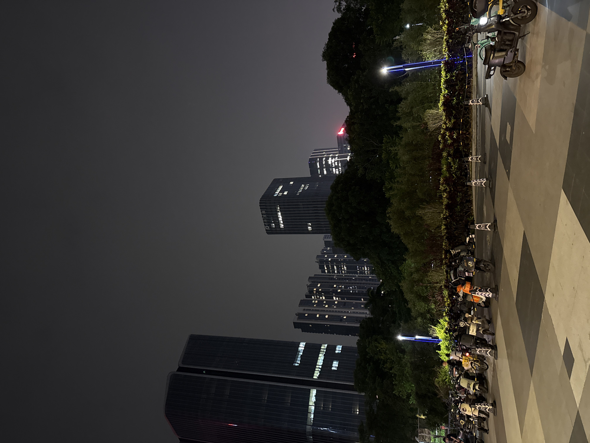 The image depicts a night scene in Huangpu District, China, focusing on a parking area filled with electric scooters and motorcycles. In the background, a row of modern high-rise buildings with illuminated windows stands against a dark sky. Several tall, dark glass skyscrapers dominate the left side of the frame, while a cluster of slightly shorter, brightly lit buildings is visible further to the right. A lush green canopy of trees separates the parking area from the buildings, with a decorative border of plants running along the edge of the parking lot.
The foreground features a checkered patterned pavement with several electric scooters and motorcycles parked in rows. Some of these vehicles are light-colored, while others are darker. Traffic cones with white and black markings are strategically placed among the parked vehicles. On the right side of the frame, a dark-colored electric scooter with its headlight on is prominently displayed. A single person is visible standing near a yellow bicycle in the middle of the parked scooters. Streetlights with blue accents illuminate parts of the scene. The overall atmosphere is urban and functional, capturing a typical evening in a busy district.