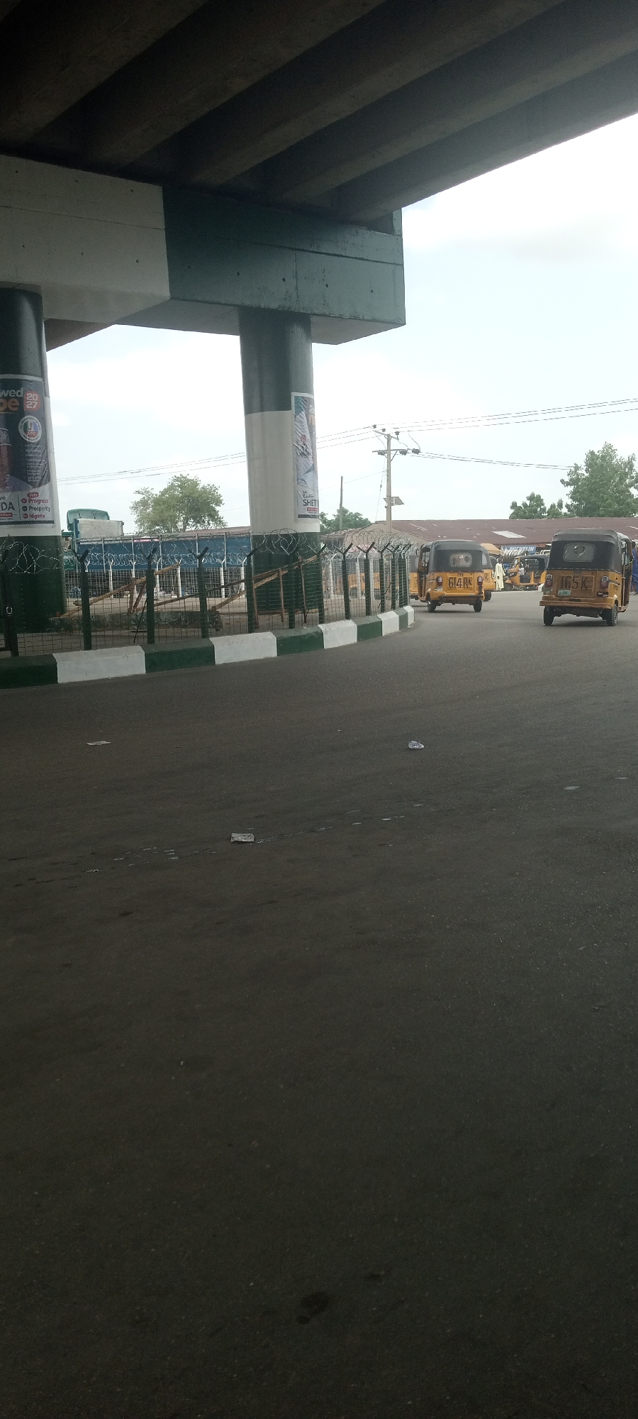 The image depicts a street scene under a large concrete overpass in Jalingo, Nigeria. The overpass supports a road, with its concrete structure visible, painted in sections of dark green and off-white. Several large, cylindrical concrete pillars, also painted in sections of dark green and white, support the overpass. Advertisements are posted on some of these pillars. A barbed wire fence runs along a sidewalk or boundary, with green and white painted concrete blocks at its base. Beyond the fence, several yellow auto-rickshaws, known as "Keke Napep" or "Keke," are parked or driving on the road. One Keke has the license plate "614 RC" and another has "165 K". In the background, there are buildings with corrugated metal roofs and some trees, suggesting an urban or semi-urban environment. The sky is overcast with a pale, cloudy appearance. The road surface in the foreground is asphalt and shows signs of litter, with several pieces of white plastic debris scattered across it. The overall scene suggests a busy transportation hub or a street corner in a Nigerian city.