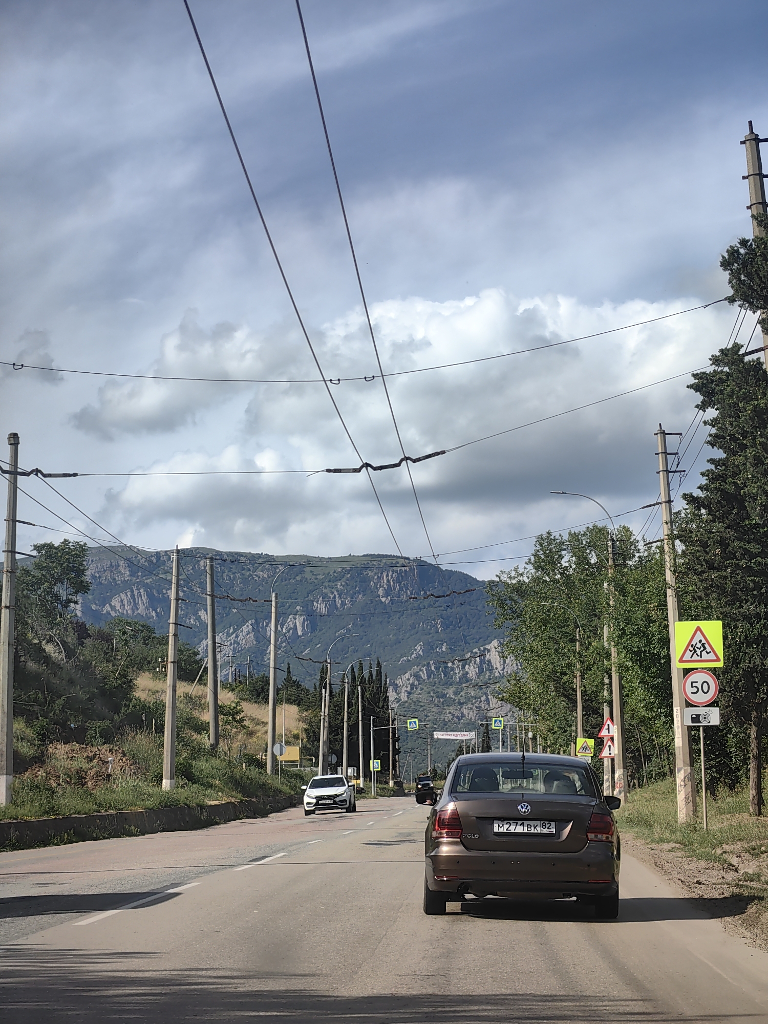 This media file captures a scenic view of a road leading towards a mountainous landscape under a cloudy sky. In the foreground, a brown Volkswagen Polo drives on a paved road. The car has a license plate that reads "M 271BK 82". A white car is visible further up the road on the left lane.
The roadside is lined with trees and utility poles supporting overhead wires. A few road signs are visible: a yellow triangular sign with figures of children running, a circular sign with the number "50" inside, and a sign with a camera icon below the speed limit sign. Another yellow triangular sign with an exclamation mark is also present.
In the background, a rugged mountain range with steep cliffs and vegetation dominates the horizon. The sky is filled with dramatic clouds, suggesting it might be a partly cloudy day. The overall impression is one of a journey through a natural, somewhat wild landscape, likely during the daytime. The location is identified as Verkhnyaya Kutuzovka, Ukraine.