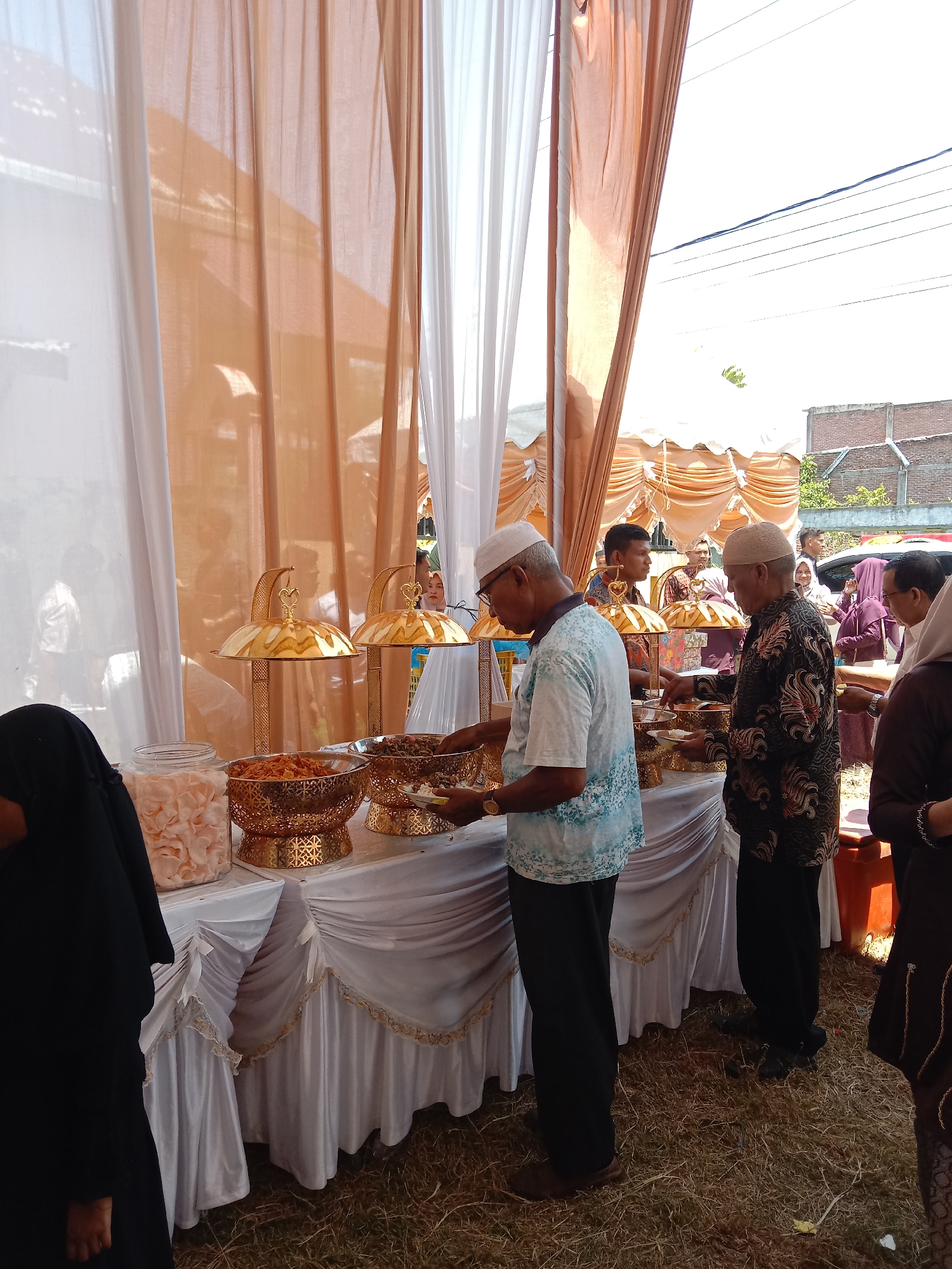 The image depicts an outdoor buffet setup in Lhokseumawe, Indonesia, likely at an event such as a wedding. The setup includes tables draped with white fabric and adorned with gold-colored serving dishes. Several people are present, many wearing traditional Muslim attire, including head coverings. They appear to be serving themselves food from the buffet. The background includes tents and structures of the event. The ground is covered with dry grass.
