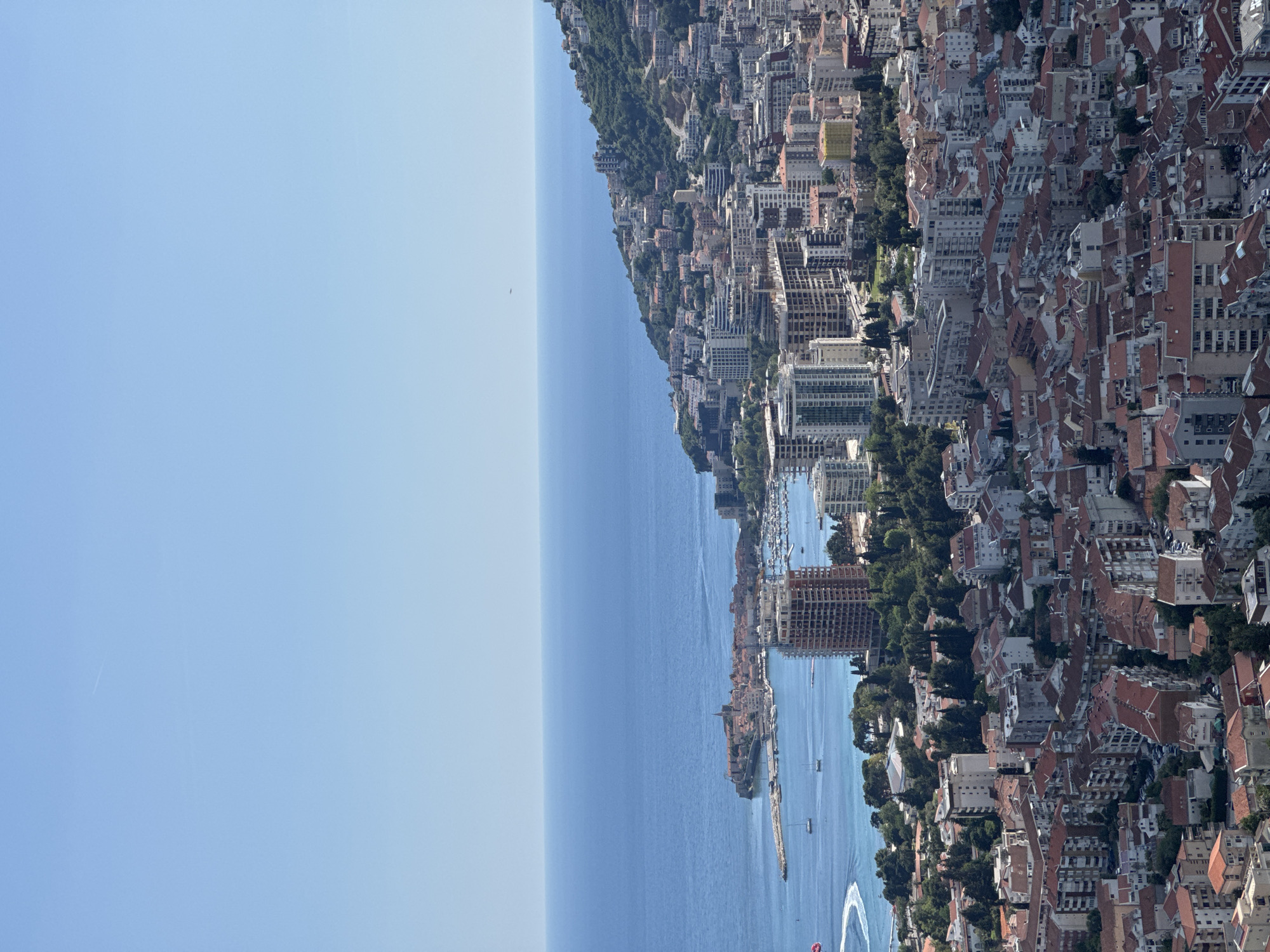 The image depicts a coastal cityscape, potentially Budva, Montenegro, with a large expanse of the Adriatic Sea on the left and the city extending to the right. The architecture consists of tightly packed buildings, many with red tile roofs, interspersed with trees and green spaces. Modern high-rise buildings and older structures are visible. Boats are seen in the water near the coastline. The sky is clear and blue.