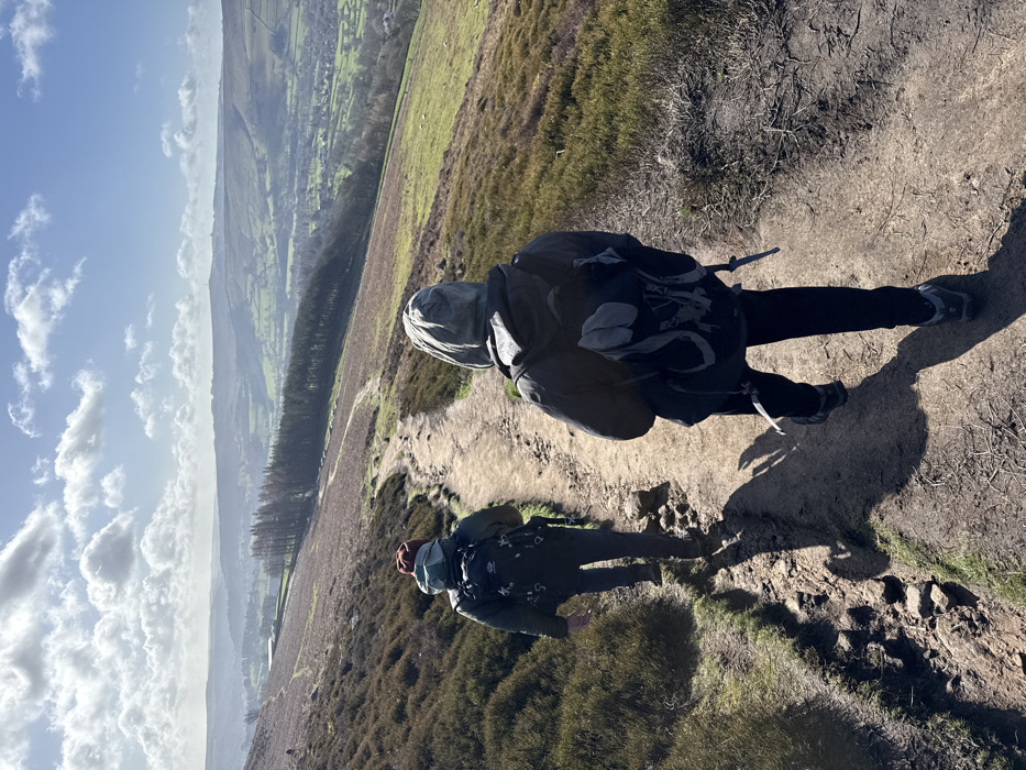 Two individuals are observed from behind, hiking along an unpaved, winding dirt path ascending a hillside within the High Peak area of the United Kingdom. The person closer to the foreground on the right wears a dark hooded jacket with the hood up, dark trousers, and a grey and black hiking backpack. The second individual, positioned further along the path on the left, wears a dark jacket, a reddish-brown beanie, and a black hiking backpack featuring light grey elements. Both are wearing hiking boots and cast distinct shadows on the path, indicating bright sunlight. The immediate hillside is predominantly covered with dry, brown heather and scattered patches of green grass. In the background, a vast valley is visible, characterized by green pastures, scattered trees including a distinct line of conifers, and distant rolling hills under a bright blue sky with numerous white cumulus clouds.