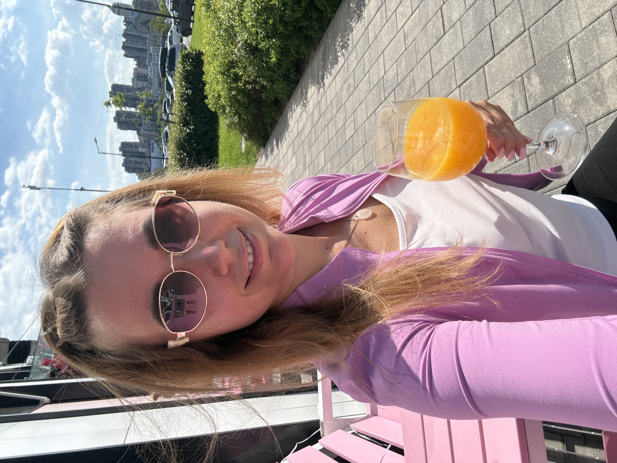 A selfie-style photo captures a woman sitting outdoors on a bright, sunny day. She has long, light brown hair, pulled back on one side, and is wearing round, gold-framed sunglasses that reflect a blurred outdoor scene. She's dressed in a white tank top layered under a light purple, long-sleeved top or jacket. Her left hand, with light pink manicured nails, holds a clear wine glass filled with a vibrant orange beverage, possibly orange juice or a cocktail. A small, clear, light pink or rose quartz pendant hangs around her neck.
The woman is smiling broadly, looking directly at the camera. She appears to be seated on a pink slatted bench or chair, visible in the foreground. The background reveals a paved sidewalk made of grey rectangular blocks. Beyond the sidewalk, there's a strip of lush green bushes and grass, followed by a street with several parked cars. In the distance, under a blue sky with fluffy white clouds, a cluster of tall, modern city buildings is visible, suggesting an urban or suburban setting. The overall scene conveys a relaxed outdoor moment, likely during the daytime, given the bright sunlight and clear shadows. There is no visible text in the image.