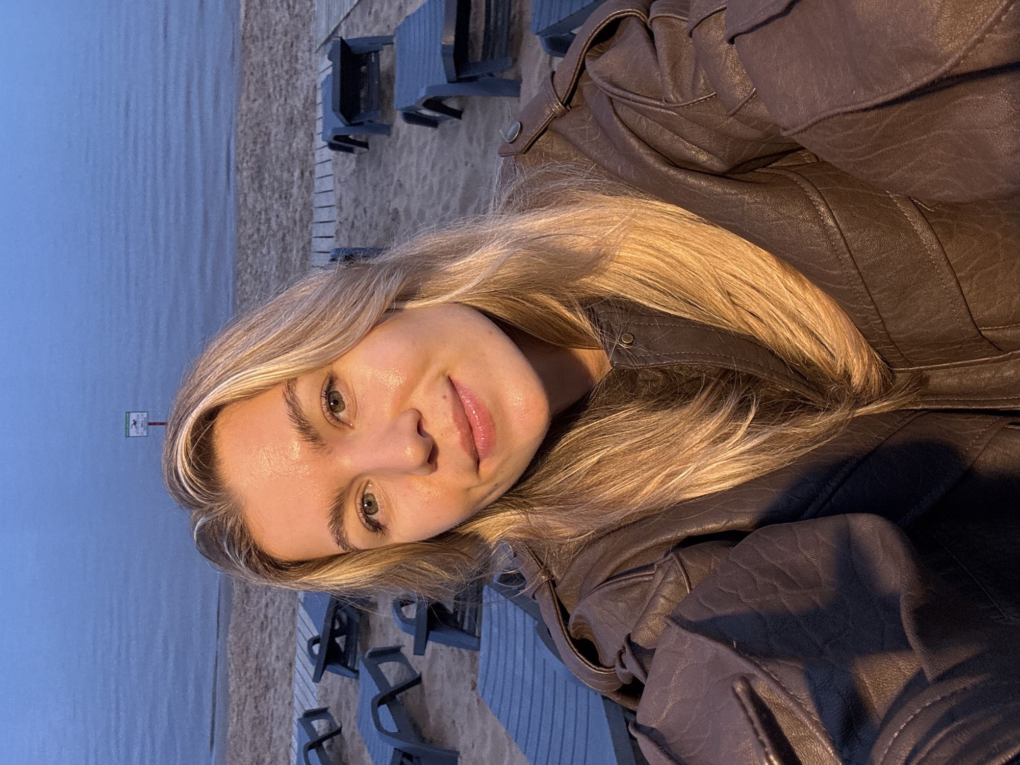 A young woman with blonde hair and green eyes smiles at the camera while wearing a brown leather jacket. She is on a beach with lounge chairs behind her and the ocean in the background. The lighting suggests it is either sunrise or sunset.