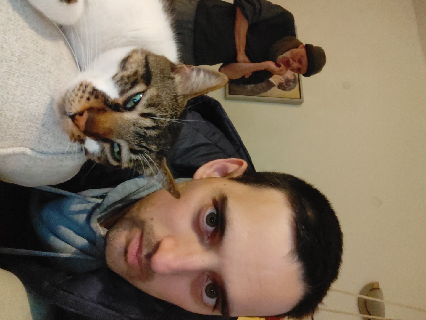 A selfie shows a fair-skinned man with dark hair and eyes, who is likely in his twenties or thirties, lying down. A white and tabby cat with green eyes is positioned above him, resting on a cushion or pillow. In the background, a painting of an older man with a beard and a cap hangs on a wall. The man in the painting is gazing towards the camera with a contemplative expression. The room appears to be indoors, and the wall is a light color. There is no indication of the location being Tel-Aviv, Israel.