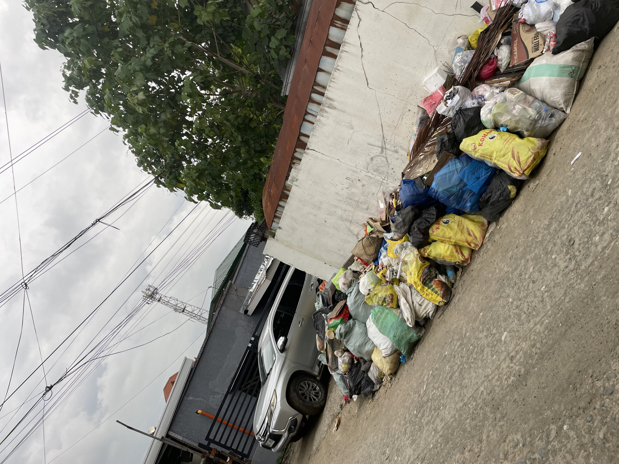 This image captures a street scene in Davao City, Philippines, depicting a significant accumulation of waste alongside a road. The setting is an urban or semi-urban environment, characterized by dense infrastructure and signs of everyday life.
On the right side of the frame, a massive pile of garbage is stacked against a light-colored, cracked concrete wall topped with a rusty corrugated metal roof or awning. The refuse consists of numerous plastic bags in various colors—yellow, blue, black, green, and white—many appearing full. Visible loose trash includes cardboard boxes, plastic bottles, and other debris. Text on some yellow bags indicates "KOHAN...", "SPECIAL RICE", and "NET WT. 25 KG", suggesting they are repurposed rice sacks. A white and green sack also reads "PREMIUM GRAINS," "JASMINE RICE," and "NET WT. 25KG." A cardboard box part of the pile displays "CAMEL" and "WALL FAN."
Adjacent to the garbage and the wall, a silver SUV, possibly a Mitsubishi Montero Sport, is parked, partially obscured by the trash. Further to the left, a dark grey building with a green roof is visible. The foreground shows a rough, grey concrete or asphalt surface forming the road or sidewalk.
Above, the sky is overcast and cloudy, indicating a daytime setting without direct sunlight. Numerous power lines and utility cables crisscross the upper left portion of the image, extending across the sky. Lush green tree foliage is visible in the top left corner, overhanging the wall. In the distance, against the cloudy sky, a tall communications or cell tower can be seen.
The scene portrays a common urban challenge of waste management, with the large, untended garbage pile being a prominent feature. No people are visible, and no specific activity or event is taking place beyond the stationary car and the accumulated waste. The overall impression is one of a busy, lived-in area grappling with environmental concerns.