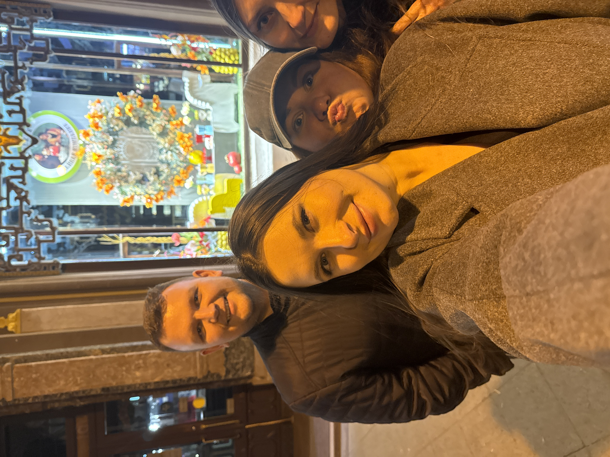 Four people are posing closely together for a selfie in front of a shop window. From left to right, there is a man in a dark jacket, a woman with long dark hair in a gray coat, a woman with a gray cap making a kissy face, and another woman. The shop window behind them displays decorative flower arrangements and what appears to be a plate with a picture. The shop front has detailed decorative trim. The location is likely Moscow, Russia, given the style of architecture and signage visible in the window.