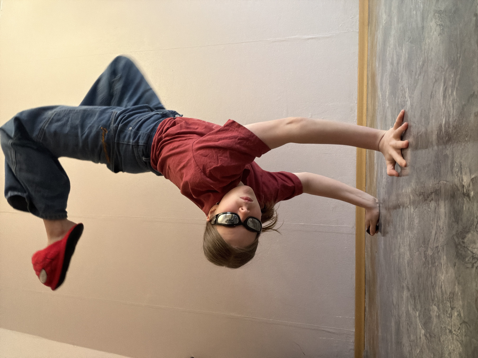 A young person is captured indoors, performing an acrobatic pose that creates an illusion of defying gravity. The image is rotated 90 degrees clockwise, presenting the person seemingly suspended horizontally, with their hands pressed against a wall.
The subject is wearing a plain red short-sleeved t-shirt, loose-fitting blue jeans, and dark, reflective sunglasses. On their visible left foot, they wear a distinctive red slipper with a white circular design and black lines, reminiscent of a Spider-Man mask. Their arms are extended towards the right, with both hands flat against a grey wall. The person's head is tilted downwards, and their face, partly obscured by sunglasses, looks directly at the camera.
The background consists of two different wall surfaces. To the left (above the person in the rotated view) is a smooth, light beige or off-white wall. To the right, where the person's hands are placed, is a textured grey wall with a mottled, possibly concrete-like finish. A narrow, golden-brown vertical strip, resembling a door frame or trim, separates these two wall sections. The lighting is soft and consistent, indicative of an indoor setting during daytime. No text is visible in the image. The overall impression is one of playful activity and creative posing. The location context for this image is Perm, Russia.