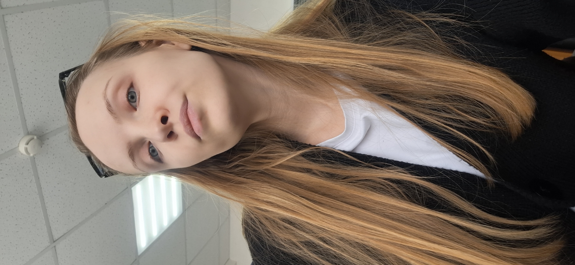 A medium close-up shows a young woman with light skin, blue eyes, and long strawberry-blonde hair. She wears a white t-shirt under a black cardigan. Sunglasses are perched on her head. The background reveals a commercial interior, presumably in a building in Chaikovsky, Russia. Details in the background include a suspended ceiling with fluorescent lights, and a white circular fire alarm.