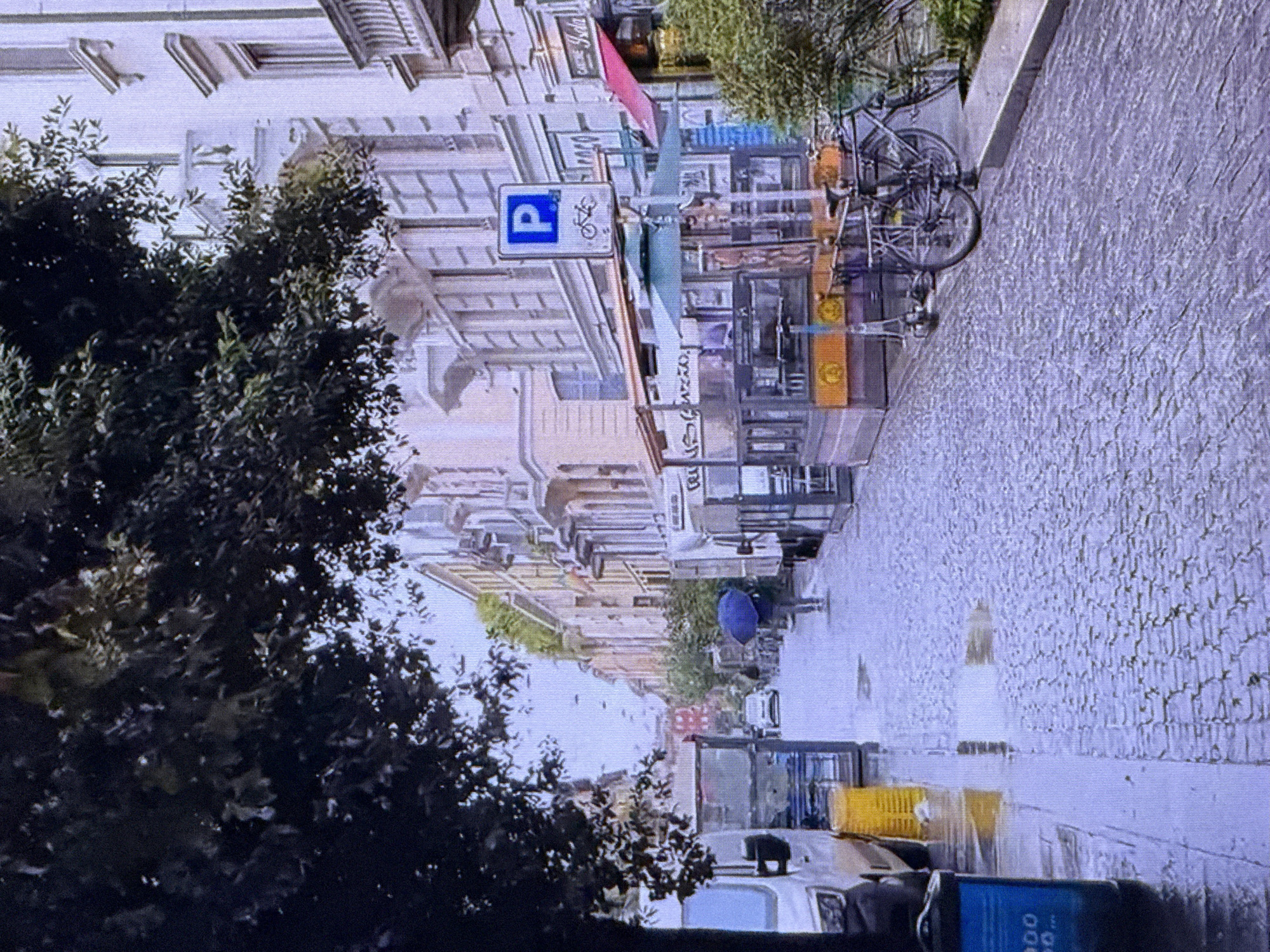 The image depicts a street scene on a rainy day. A person holding a blue umbrella is walking down the cobblestone street. The street is lined with buildings, some of which have shops or cafes with outdoor seating areas covered by awnings. There are vehicles present, including a white van on the left side of the street and a car further down. The environment appears wet, with reflections visible on the cobblestones, suggesting recent rainfall. The trees overhang the street, with some foliage visible. A parking sign with a bicycle symbol is visible on a pole. There is a bicycle and a scooter parked near the shops. The overall impression is of an urban environment experiencing inclement weather. The image does not show any indication of being in Dubai, United Arab Emirates; rather, the architecture and streetscape suggest a European city.