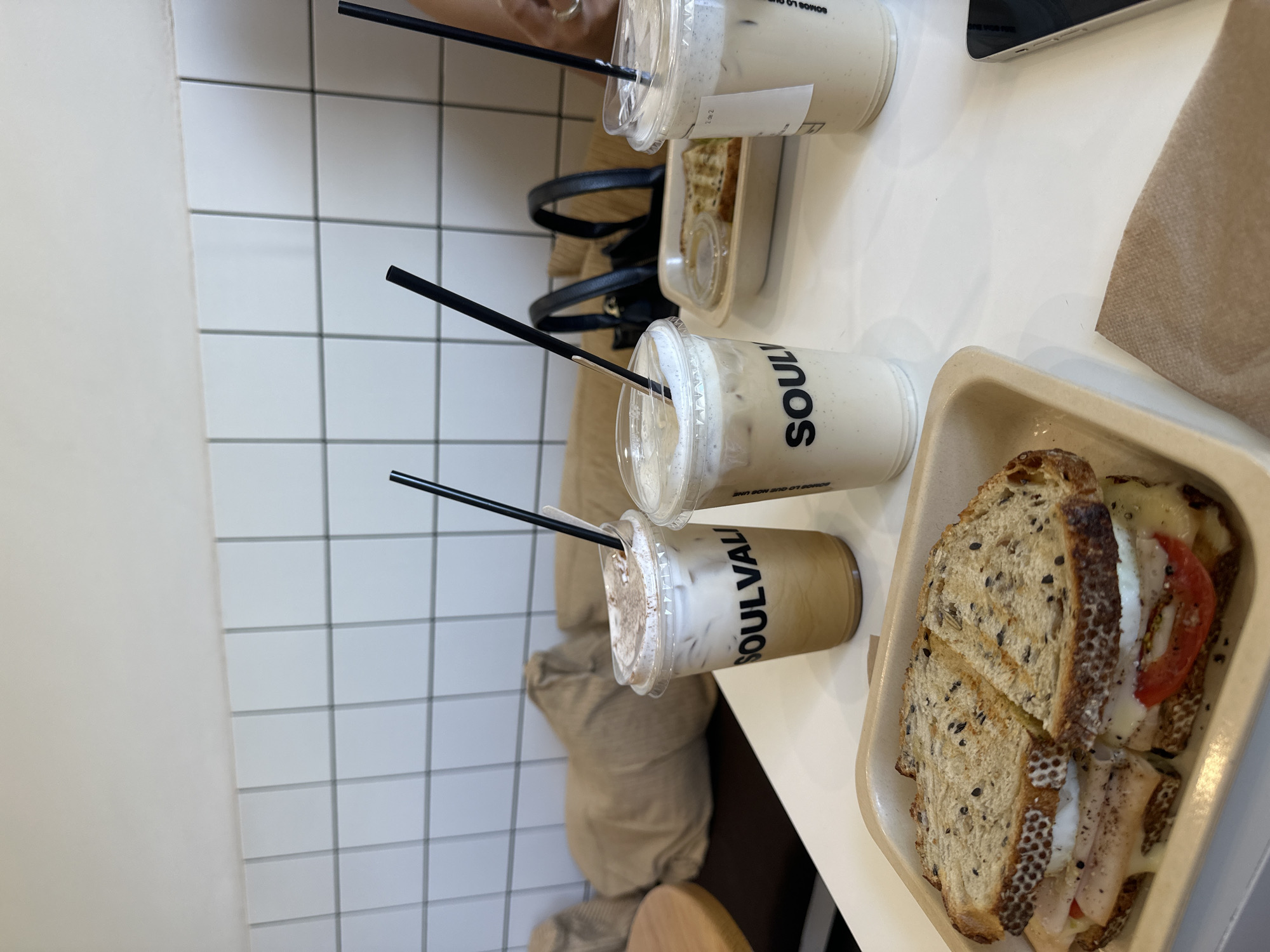 The image depicts an indoor scene at Soulvali Café, located at 3 Sur 704, Centro Histórico, Puebla, Mexico. A white tabletop serves as the primary surface. Three clear plastic cups containing iced beverages with foamed tops and black straws are present. Two cups clearly display the brand name "SOULVALI".
In the foreground, a multi-grain sandwich, cut diagonally into two halves, rests on a light brown rectangular tray. The sandwich filling consists of sliced deli meat (likely turkey), melted yellow cheese, red tomato slices, and a white, possibly egg-based, component, all seasoned with black pepper. A brown paper napkin is visible directly beneath the sandwich tray.
In the mid-ground, a smaller light brown container holds two pieces of toasted bread and a small, lidded clear plastic cup containing a condiment. A black handbag with two top handles is situated behind these items. A person's arm and hand are partially visible on the right side of the frame, adjacent to the third beverage. A dark-colored smartphone is partially visible on the table in the lower right corner.
The background features a wall covered with white square tiles. Below the tiled wall, a seating area with dark brown cushions and large, light brown fabric pillows is visible. A circular wooden table edge is partially visible on the far left. The lighting is bright and even.