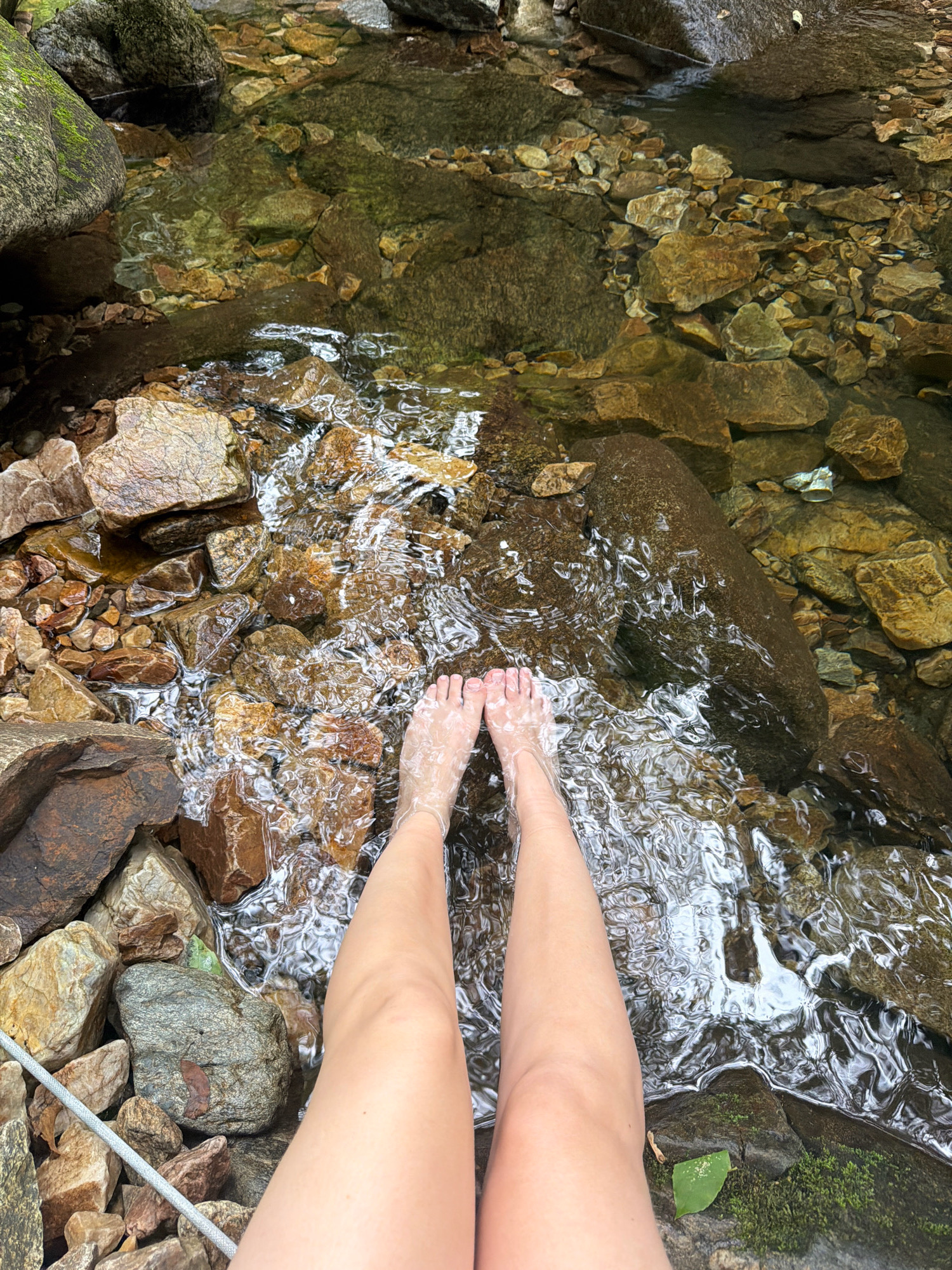 The image shows a pair of human legs and feet submerged in clear, shallow water flowing over a rocky stream bed. The water is slightly rippled, reflecting the surrounding environment. The stream bed is composed of various sizes of rocks, some appearing wet and darker, while others are lighter and drier. Green moss is visible on some of the larger rocks, particularly on the left side of the frame. The surrounding area appears to be a natural, outdoor environment with rocks and possibly vegetation. The perspective is from above, looking down at the legs and feet. The image does not provide any specific clues that would place it in Dongducheon-si, South Korea.