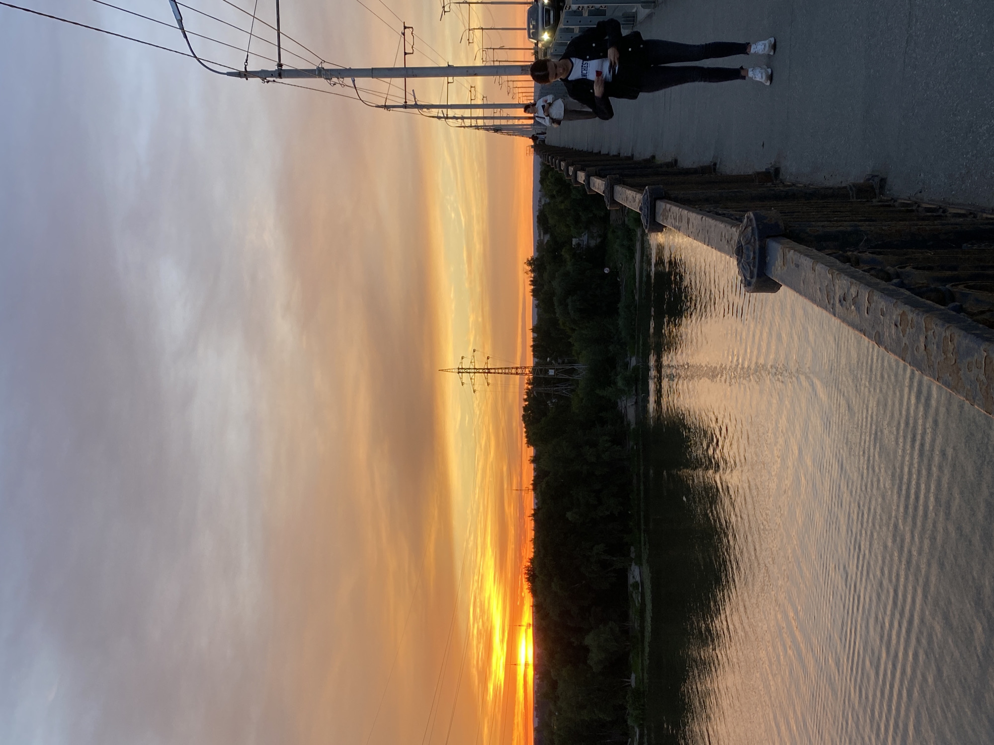 This is a photograph taken from a bridge over a river in Novokuybyshevsk, Russia, during sunset. The sky is filled with a vibrant orange and yellow glow, with streaks of pale blue and gray clouds. On the left side of the frame, lush green trees line the riverbank, with reflections of the sunset visible on the water's surface. The bridge railing, made of dark metal with decorative elements, runs along the right side of the image. A young woman with dark hair, wearing a black jacket over a white t-shirt and dark pants, is walking on the bridge, looking down at her phone. Another person is visible further down the bridge, as well as a car driving in the distance. Electrical poles and wires stretch across the top of the frame, indicative of urban infrastructure. The overall scene captures a serene evening atmosphere.
