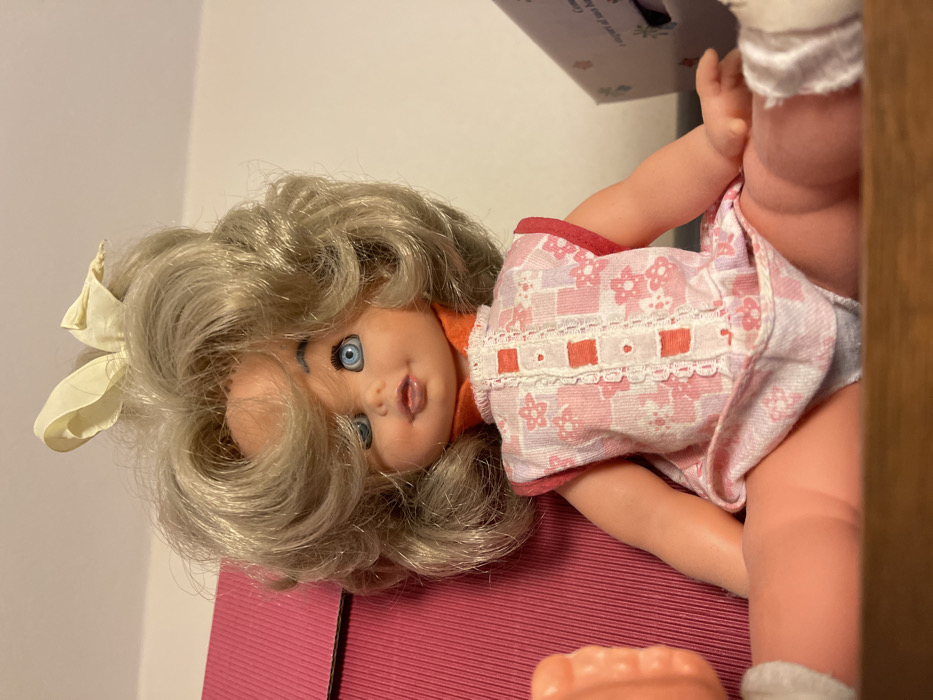 An indoor, brightly lit close-up shot captures a doll with blonde, curly hair and striking blue eyes. The doll's head is positioned towards the left of the frame, lying on its back, with its mouth slightly open, revealing a glimpse of its tongue. A light yellow bow is tied in its hair on the left side. The doll wears a light pink and white patterned dress, possibly featuring floral or geometric designs, accented with a white lace trim across the bodice. An orange garment appears to be worn underneath the dress, visible around the doll's neck.

The doll is resting on a reddish-pink, ribbed surface, which could be a mat or a box lid. In the upper right background, a blurred, light-colored object, possibly a box, displays faint, illegible text. A brown wooden surface frames the far right edge of the image. Part of another doll's or the same doll's leg and foot, wearing a white sock, is visible on the right side of the frame. The setting is ambiguous but appears to be indoors.