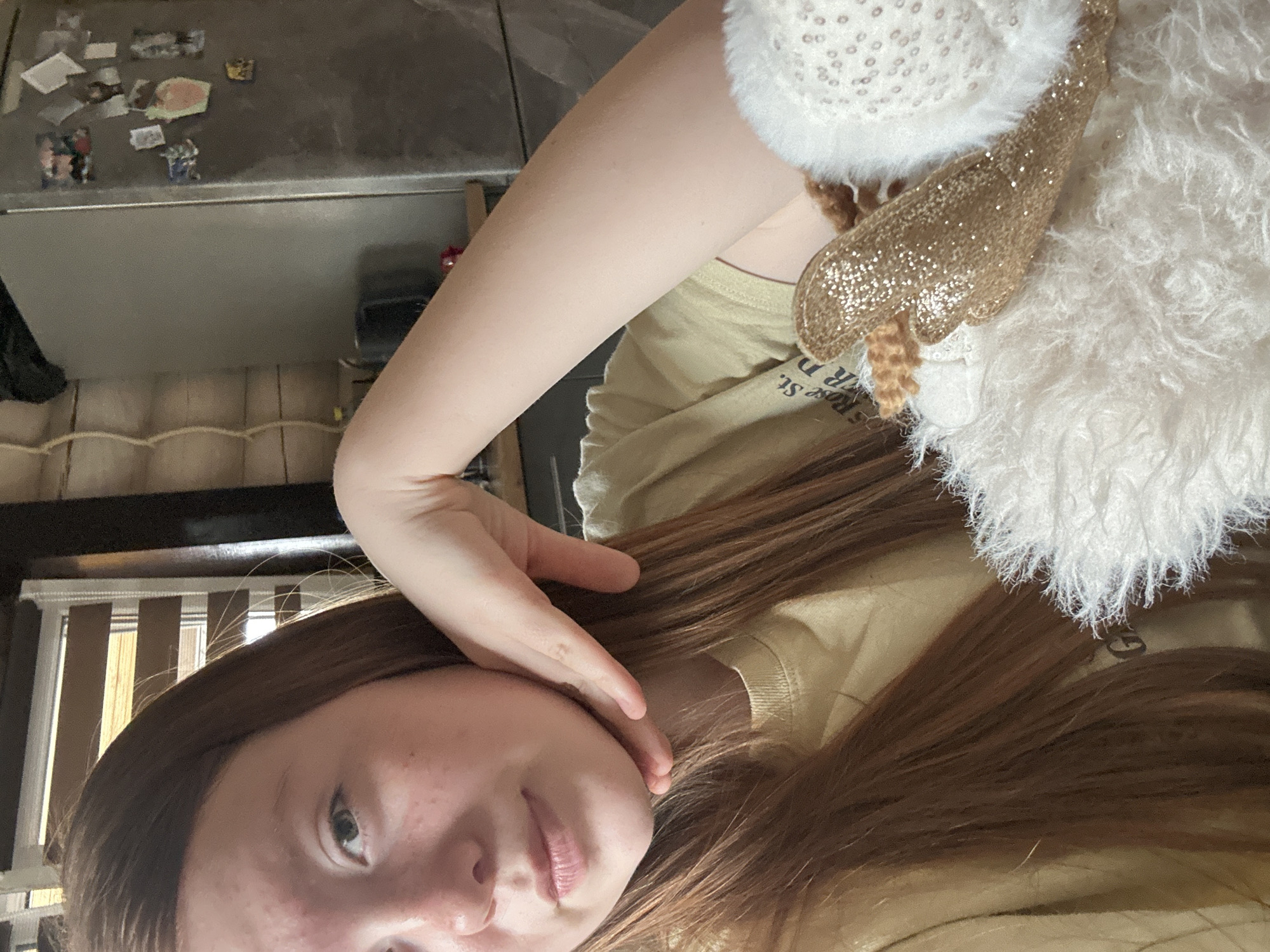 A girl is lying down in front of a window with blinds, wearing a light-colored t-shirt and a white fluffy accessory with a gold-colored accent. Her hand is touching her face. Behind her is a refrigerator with items attached to it, and wood paneling. There is no indication that the location is in the city of Ольховка, Russia.