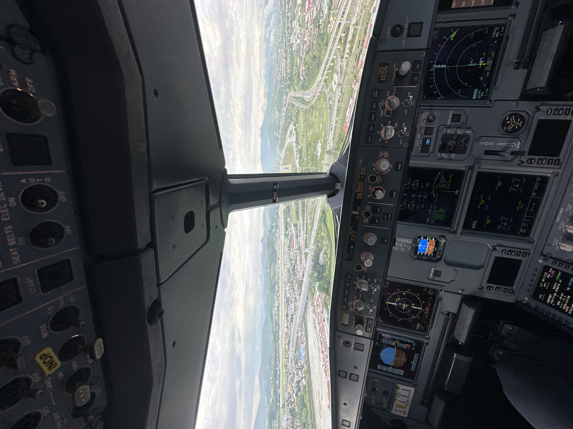 The image shows a cockpit view, likely from a flight simulator, with a clear view of a city that resembles Sochi, Russia, during daylight. The plane appears to be approaching or departing, with the city sprawling beneath. Multiple highways and structures are visible.
The cockpit instruments are prominently displayed. Visible readouts include "06000 -3.0", "1006", and "14.5240". Various buttons and knobs are present.