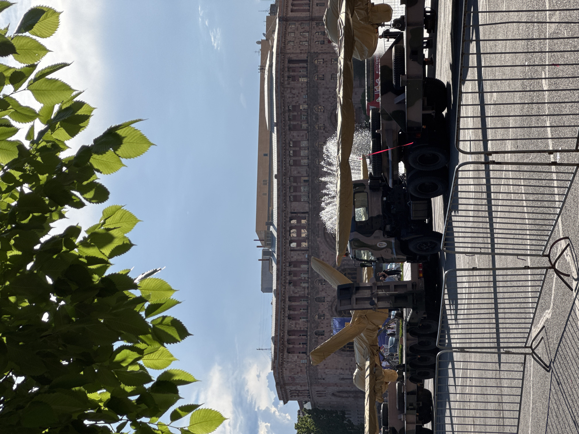 The image depicts a street scene in Yerevan, Armenia, featuring military vehicles lined up along a street. A large building, likely located on Republic Square, is visible in the background. Metal barricades run along the edge of the street, and green foliage frames the left side of the image. Water is being sprayed from one of the vehicles onto the building in the background. The weather appears sunny.