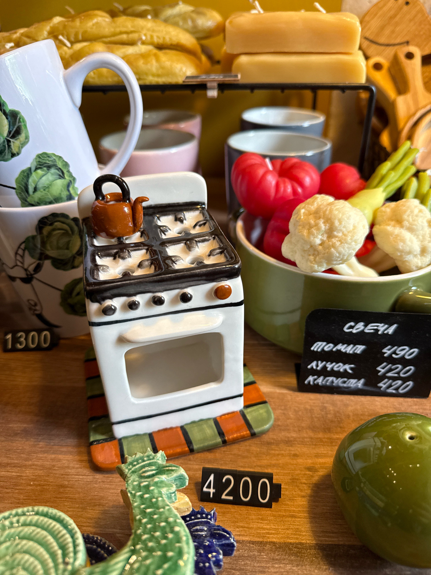 In Kaliningrad, Russia, on a wooden surface, a collection of novelty candles are displayed. In the foreground, a miniature white stove candle holder, adorned with a tiny orange teapot on top, sits on a colorful mat. To the right, a black chalkboard lists prices for various candle scents: "СВЕЧА" (Candle) with "ТОМАТ" (Tomato) at 490, "ЛУЧОК" (Onion) at 420, and "КАПУСТА" (Cabbage) at 420. Nearby, a shiny green ceramic olive-shaped candle is visible. Behind the stove, a green bowl holds red tomato-shaped candles and cauliflower-shaped candles. Further back, several gray ceramic cups are stacked. A white pitcher with a cabbage motif stands to the left. In the background, a rack holds several yellow, rectangular candles stacked two high. To the right of the rack, a small wooden cutting board with a carved face is present. A black price tag with "1300" is in the lower left, and another with "4200" is in the lower right. A decorative green rooster and blue crown figures are also present. The lighting suggests an indoor environment, likely a shop or market stall.
