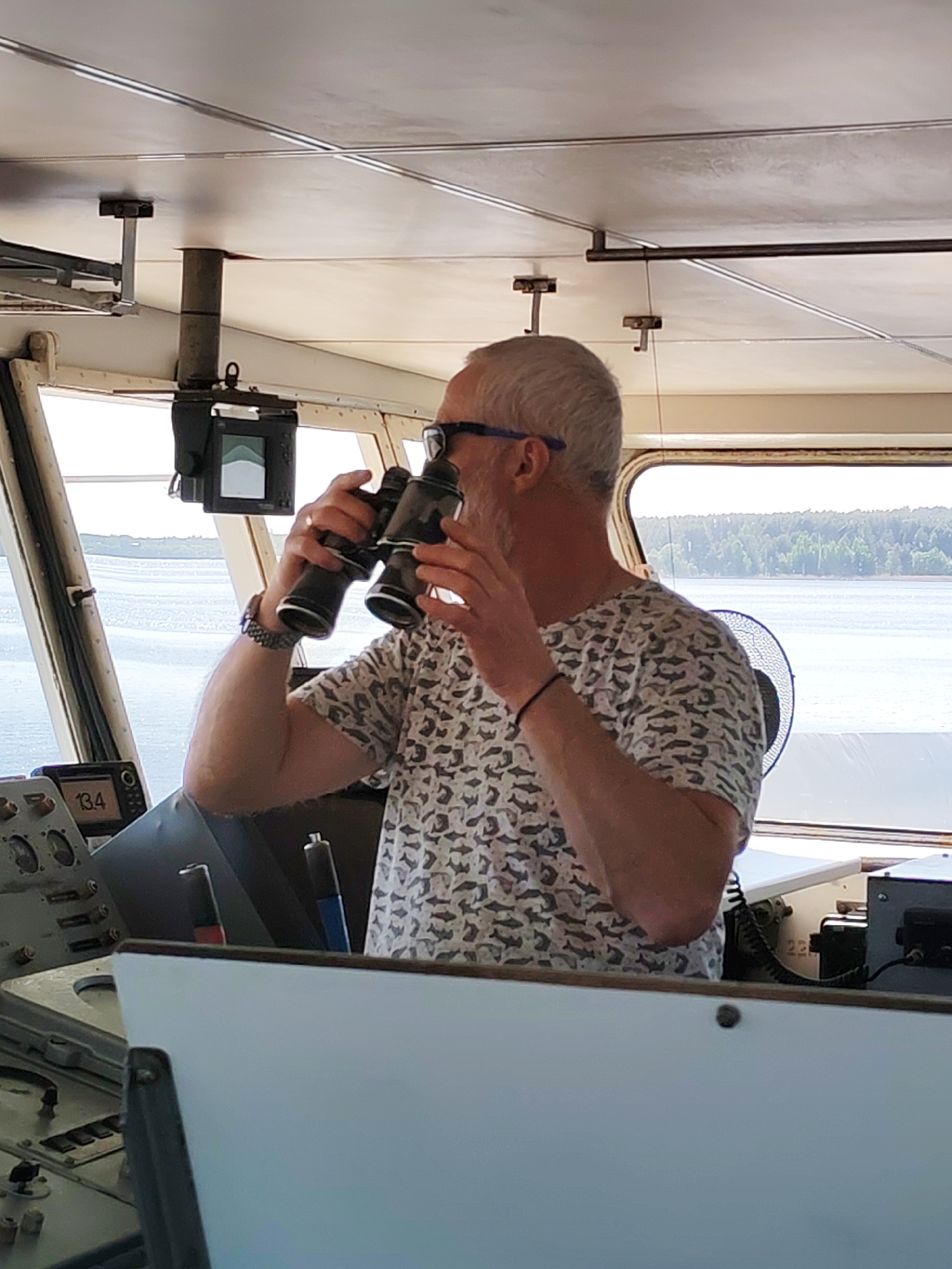 A man is inside what appears to be the bridge of a boat or ship, looking through binoculars. The man is white, with gray hair, wearing glasses and a patterned shirt. He has a watch on his left wrist and a dark bracelet on his right. He is positioned in front of a control panel with various gauges, screens, and controls. Outside the windows, there is a body of water, possibly a lake or river, with forested land visible in the distance. The interior has a utilitarian feel, with metal framing and fixtures. The specific location in Tarchevo, Russia cannot be determined from this image.