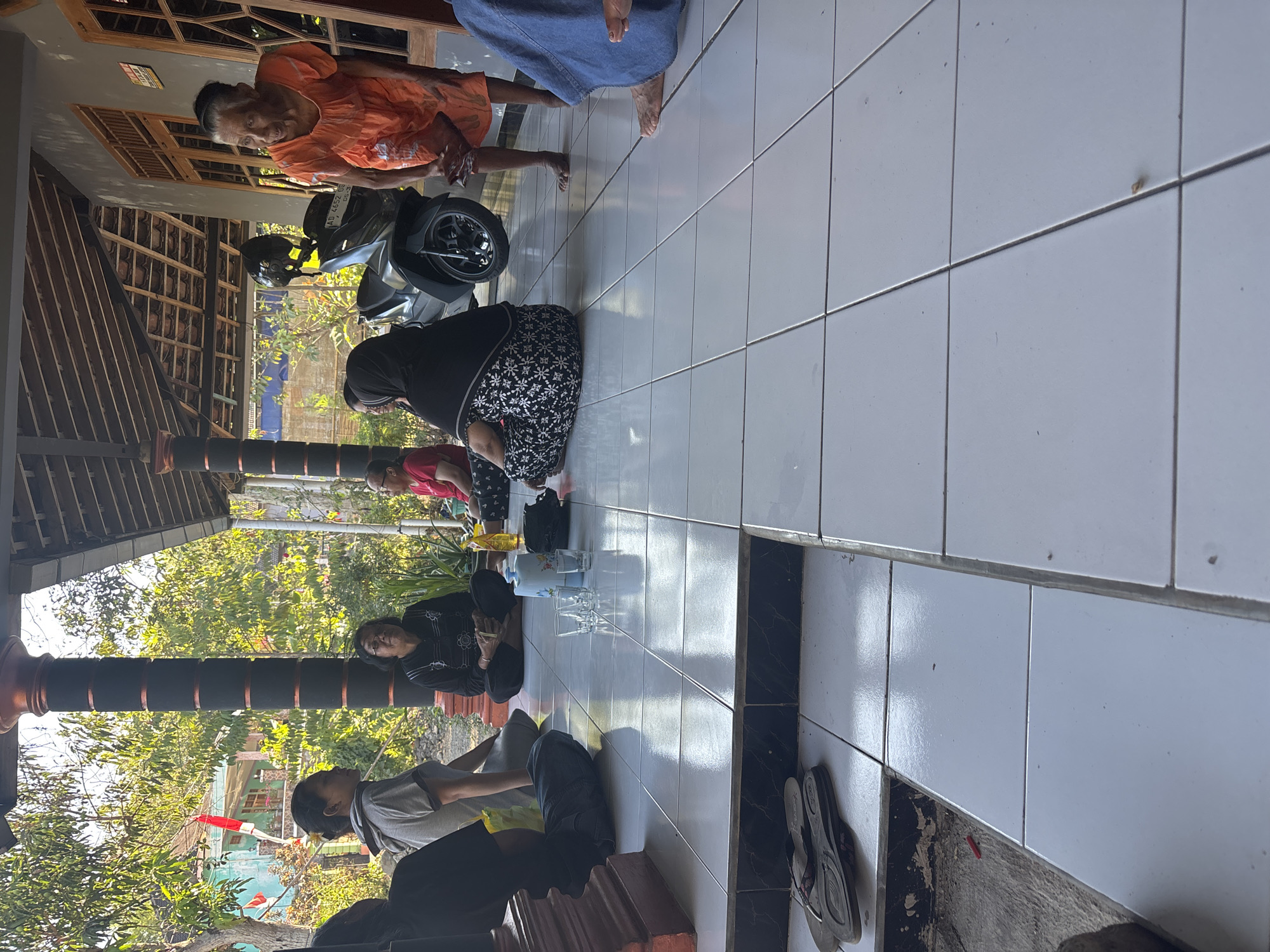 The image captures a gathering of at least six people sitting on the tiled floor of a covered porch area, likely in Tirtomoyo, Indonesia, given the context. The setting appears to be a residential area with a house visible in the background, featuring wooden window frames and a tiled roof. Lush green trees and vegetation surround the property, and a red and white flag is displayed on a nearby building, suggesting a cultural or national celebration.
In the foreground, a group is seated, engaged in conversation or simply relaxing. One individual, an elderly woman in an orange dress, stands and gestures with her hands, appearing to be speaking. To her left, a person in a black and white patterned outfit is seated, facing away from the camera. Another individual in a black headscarf is also seated. Further to the left, a person wearing a grey t-shirt and dark pants is seated, with their back to the camera. Two other individuals are partially visible, one in a red shirt and another in dark clothing.
The porch floor is tiled in a grid pattern, and a few glasses and a thermos are placed on the floor, indicating that drinks may have been served. A dark-colored motorcycle is parked behind the seated individuals. Near the edge of the porch, a pair of flip-flops is placed on a step. The overall atmosphere suggests a casual, informal gathering in a warm, outdoor environment. The lighting indicates it is daytime, likely morning or afternoon.