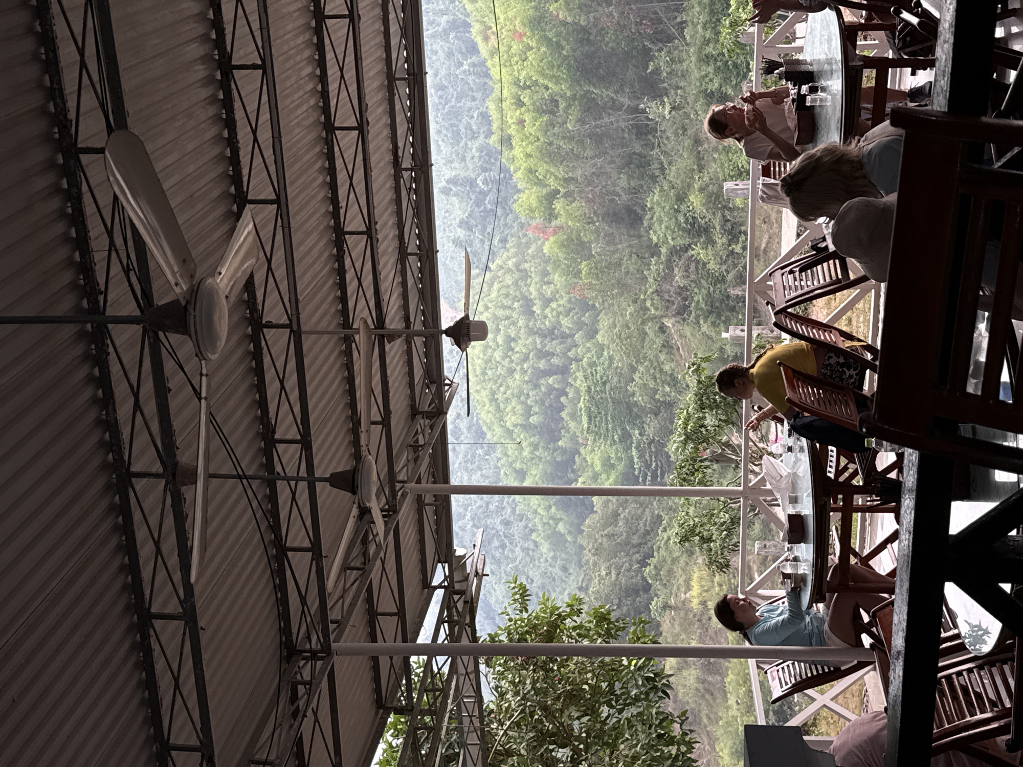 An open-air dining area at a restaurant or cafe overlooks a panoramic, misty mountain landscape in Nam Khanh Vinh, Vietnam. The establishment features a corrugated metal roof supported by dark metal trusses and several industrial-style ceiling fans. Grey support poles and a white wooden railing define the edge of the dining space.
Multiple patrons are seated at wooden tables with glass tops. In the foreground, a woman in a light blue top is seen holding a drink, seemingly relaxed. Further back, another woman with glasses is actively eating from a bowl or plate. Other individuals are partially visible at nearby tables, engaged in dining or conversation. The overall activity suggests a leisurely meal or refreshment break.
The background is dominated by a vast expanse of dense, lush green forest covering rolling mountains, receding into a soft haze or mist, characteristic of a humid climate. The time of day appears to be daytime, possibly early morning or late afternoon, given the diffused light. The scene evokes a calm and tranquil atmosphere, with people enjoying the scenic view. No visible text is present in the image.