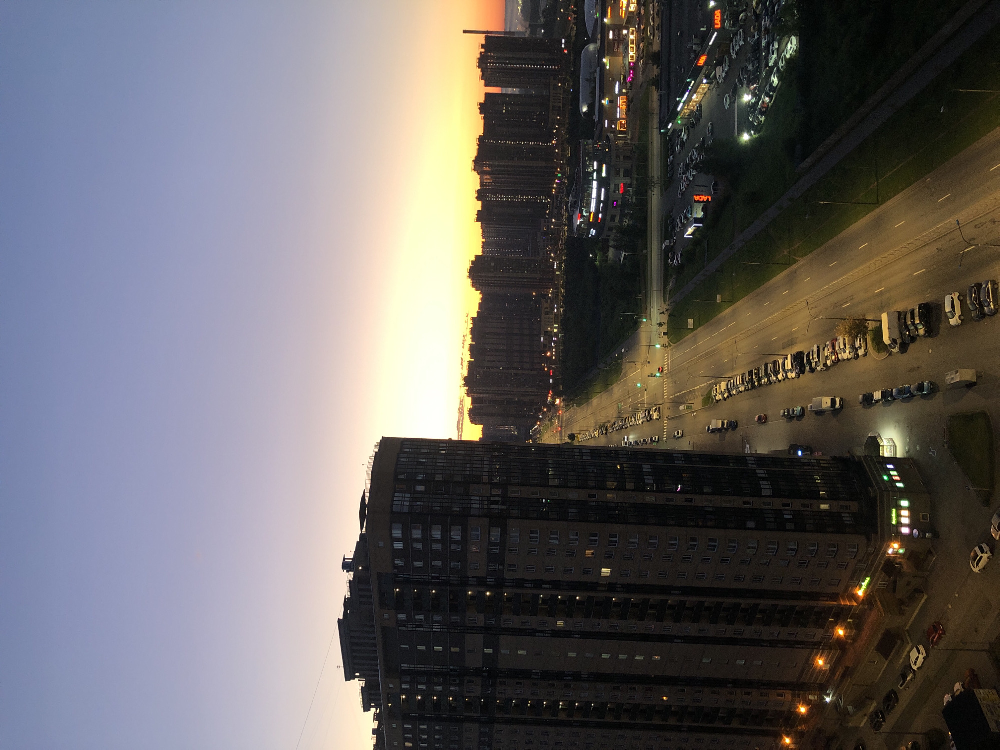 The image depicts an aerial view of a city at dusk. The sky transitions from a deep purple at the top to a bright yellow and orange horizon. In the foreground, a tall, dark-colored residential building with many illuminated windows dominates the left side of the frame. Adjacent to the building, a multi-lane road stretches into the distance, lined with parked cars on both sides. Further down the road, numerous other residential buildings are visible, forming a dense urban landscape. A few commercial buildings with illuminated signage are also present. The scene appears to be a typical urban environment during the evening transition from daylight to night.