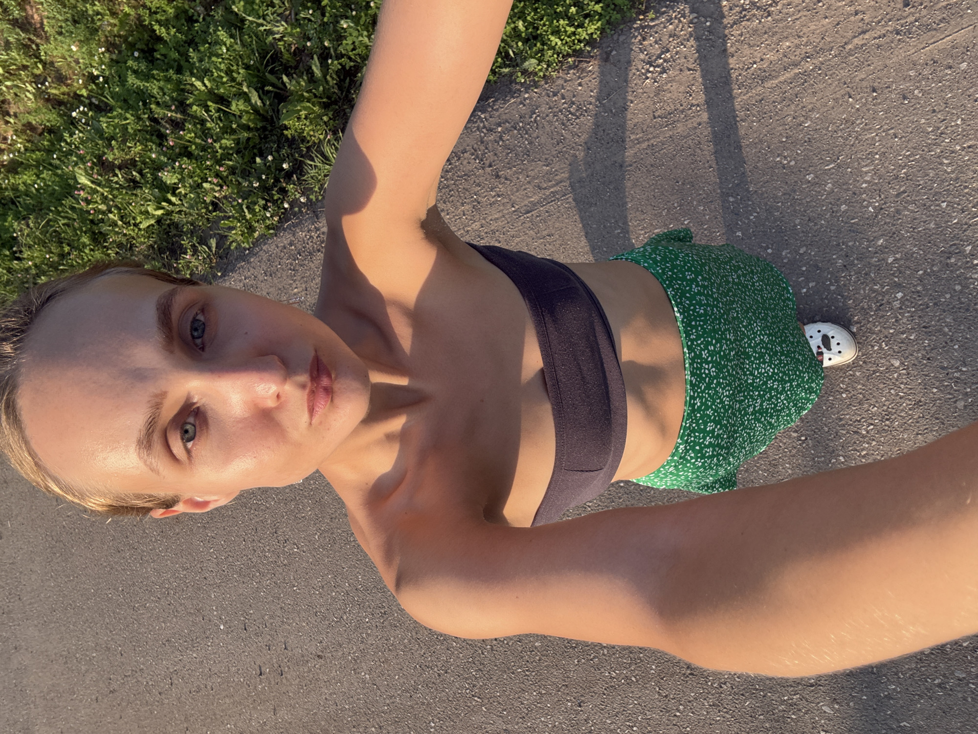 A woman with blonde hair is taking a selfie outdoors. She is wearing a dark sports bra and a green skirt with a white floral pattern. She is standing on a paved surface, possibly a road or sidewalk, with grass and some small white flowers visible to her left. The sun is casting shadows, indicating it is either morning or late afternoon. The lighting suggests a warm, sunny day. Her expression is neutral, and she is looking towards the camera. The image is taken from a high angle, looking down at the woman. There is no visible text in the image. The location is identified as Podshivalovo, Russia.
