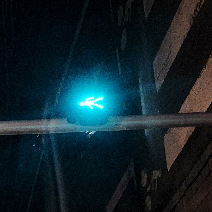 A green pedestrian crossing light, depicting a walking figure, is illuminated against a dark background. A horizontal gray bar bisects the image below the light. A zebra crossing pattern and part of the wall are visible to the right of the pedestrian light. The overall scene is dimly lit, with the light source concentrated on the pedestrian symbol, indicating it is safe to cross.