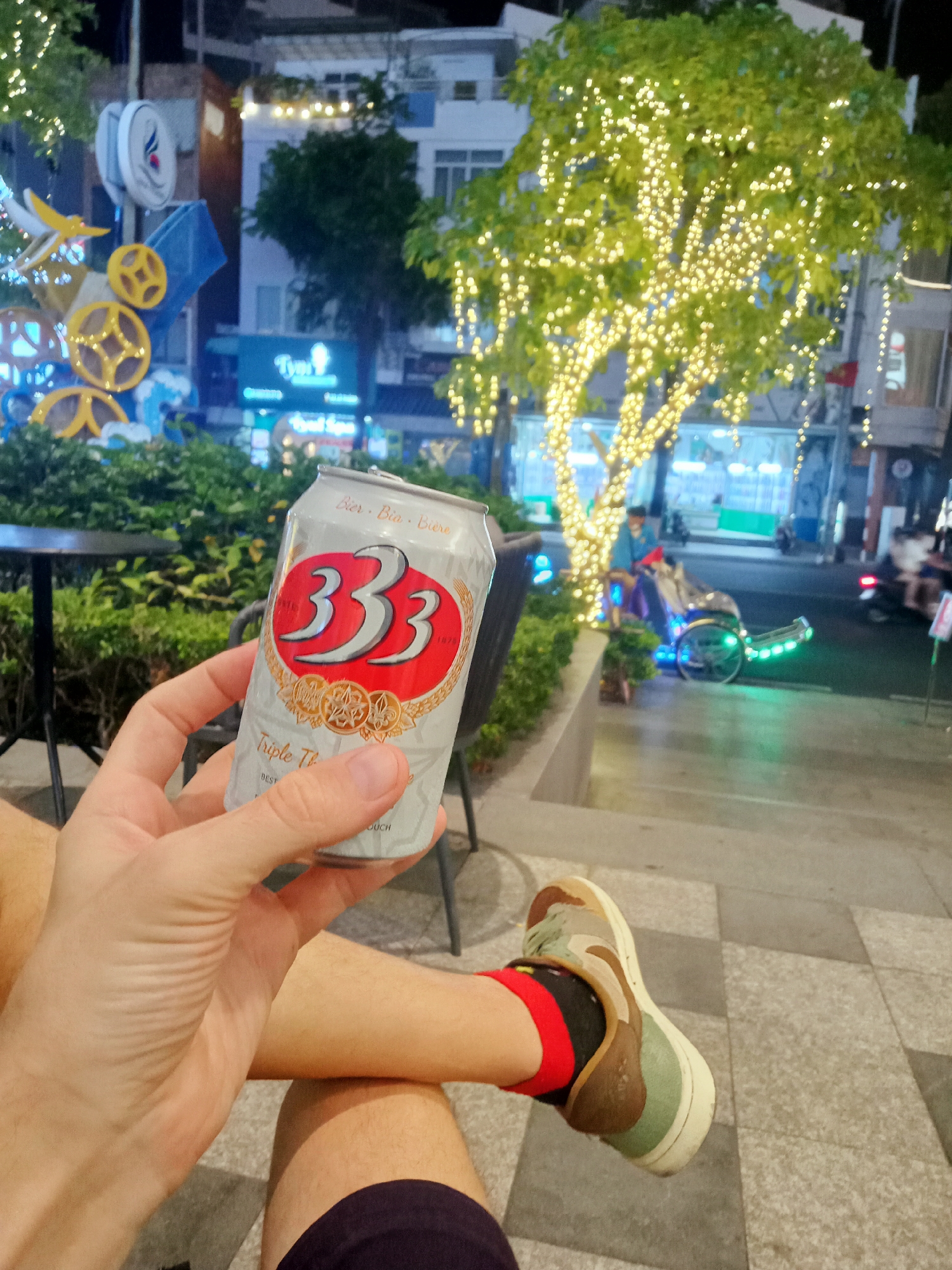 A person sits on a paved outdoor patio in Nha Trang Ward, Vietnam. They are holding a can of 333 beer. The background shows a street scene at night with buildings, decorative lights, and a lighted cycle rickshaw. The individual is wearing a black and red sock and a sneaker.