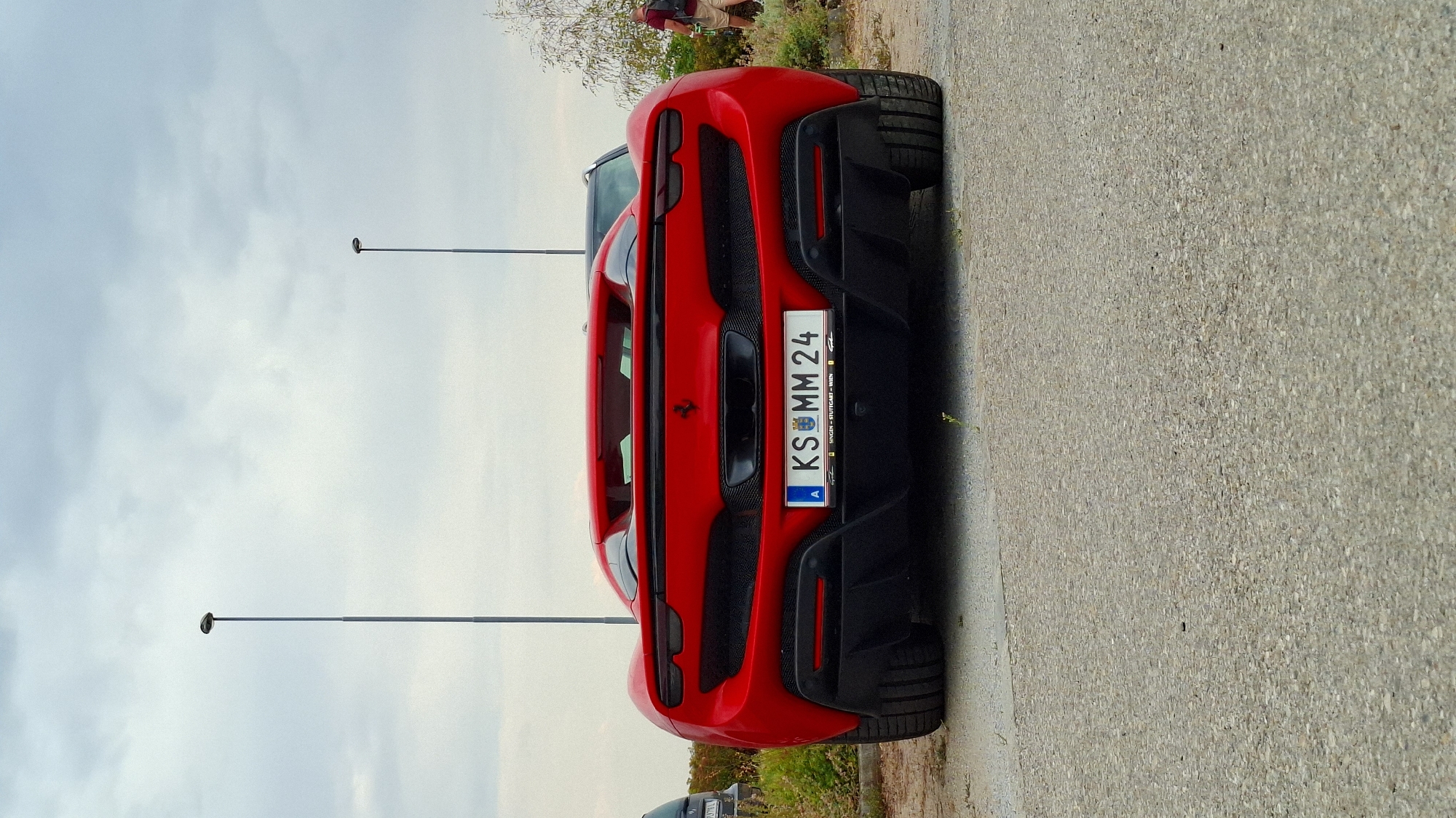 A red Ferrari sports car is parked on asphalt. The rear of the vehicle is visible, showing its license plate which reads "KS MM 24". The car has a black diffuser and black mesh on the rear. In the background, two lampposts are visible against a cloudy sky. To the right, a person wearing a red shirt and shorts stands with their back to the camera. A tree with sparse leaves is behind the person. The ground is a mix of asphalt and gravel.