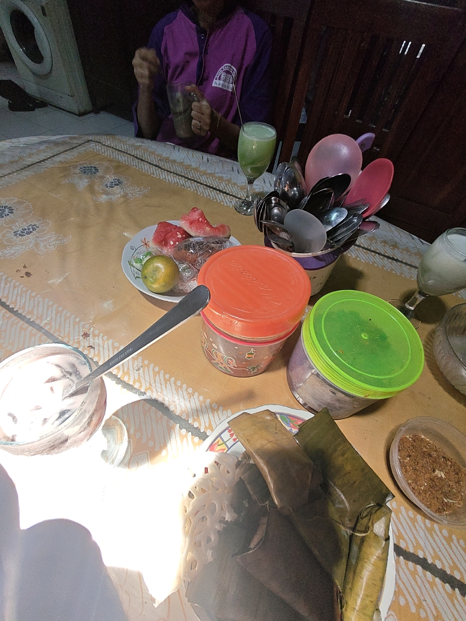 The image depicts a dining table in what appears to be a home in Sumberwono, Indonesia. A person, partially visible and wearing a purple shirt with a logo, is seated and holding a glass of green liquid. On the table, a yellow patterned tablecloth is laid out. A plate contains several slices of watermelon, an orange, and some wrapped food items. Two plastic containers, one with an orange lid and another with a green lid, are present. A bowl containing a white, creamy substance and a spoon is also on the table, along with a cluster of cutlery in a container. A pink balloon is visible near the cutlery. In the background, wooden chairs and part of a washing machine are discernible. The overall scene suggests a meal or snack time in a casual domestic setting.