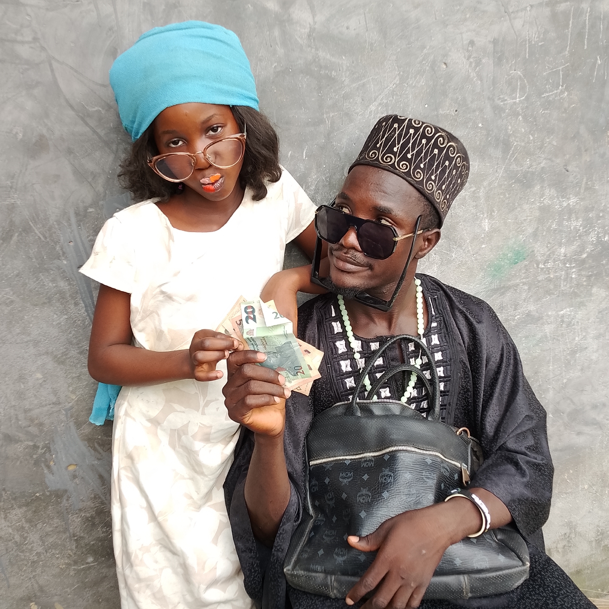 In Ado, Nigeria, a young girl wearing a blue headscarf, a white dress with floral patterns, and oversized glasses with bright orange lipstick stands leaning against a grey, textured wall. Beside her, a man in a black traditional outfit, a black cap adorned with gold embroidery, and large sunglasses holds several Nigerian Naira bills in his hand. He also wears a necklace of light green beads. A black MCM-patterned handbag rests on his lap. The background is a simple, unadorned grey wall.
