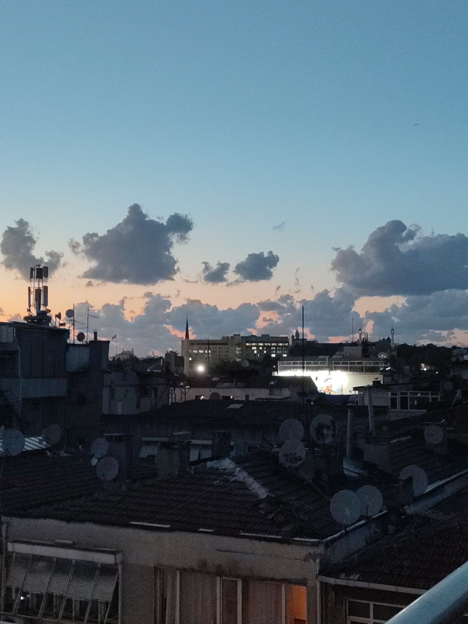 This is a vertical, eye-level, medium shot of a city skyline at dusk, with a focus on buildings and the sky. The sky is a gradient of light blue at the top, transitioning to a soft orange and pink hue near the horizon, with scattered grey clouds. In the foreground, several rooftops are visible, covered with dark, weathered tiles. Numerous satellite dishes are mounted on these rooftops.
Prominently featured on the left side of the image is a tall telecommunications tower with multiple antennae. In the mid-ground, various buildings are silhouetted against the twilight sky. Some of these buildings have lit windows, suggesting activity within. One taller building in the center features a distinct spire. The overall impression is one of a densely populated urban environment, likely residential, seen from an elevated perspective during the transitional period between day and night. The location is identified as Kadıköy, Turkey.
