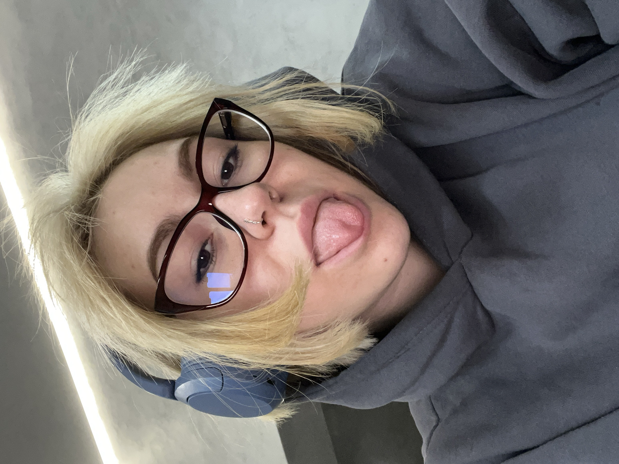 A woman with light blonde hair and glasses is positioned at an angle facing the viewer. She is wearing a gray hoodie and has headphones around her neck. Her tongue is sticking out in a playful gesture. A nose ring is visible in her left nostril. The lighting appears to be from an overhead source. It is not possible to determine the location as Odesa, Ukraine from the provided image. The environment consists of a gray surface behind her.
