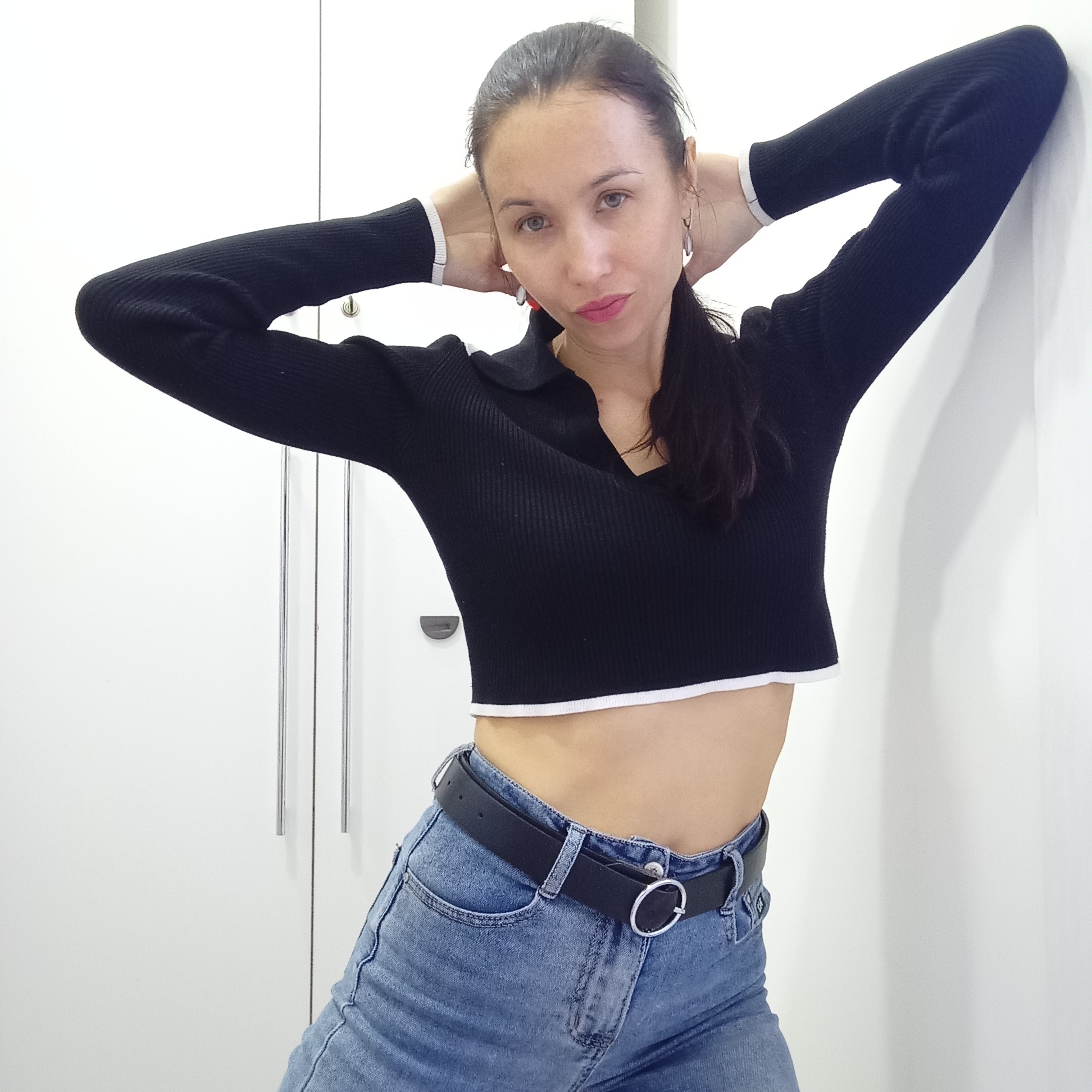 A slender woman with dark hair tied back is posing indoors. She is wearing a black, long-sleeved, ribbed crop top with white trim at the cuffs and hem, and blue denim jeans with a black leather belt featuring a silver circular buckle. Her arms are raised behind her head, and she leans against a white wall next to a white wardrobe with vertical silver handles. The location is not identifiable from the image.