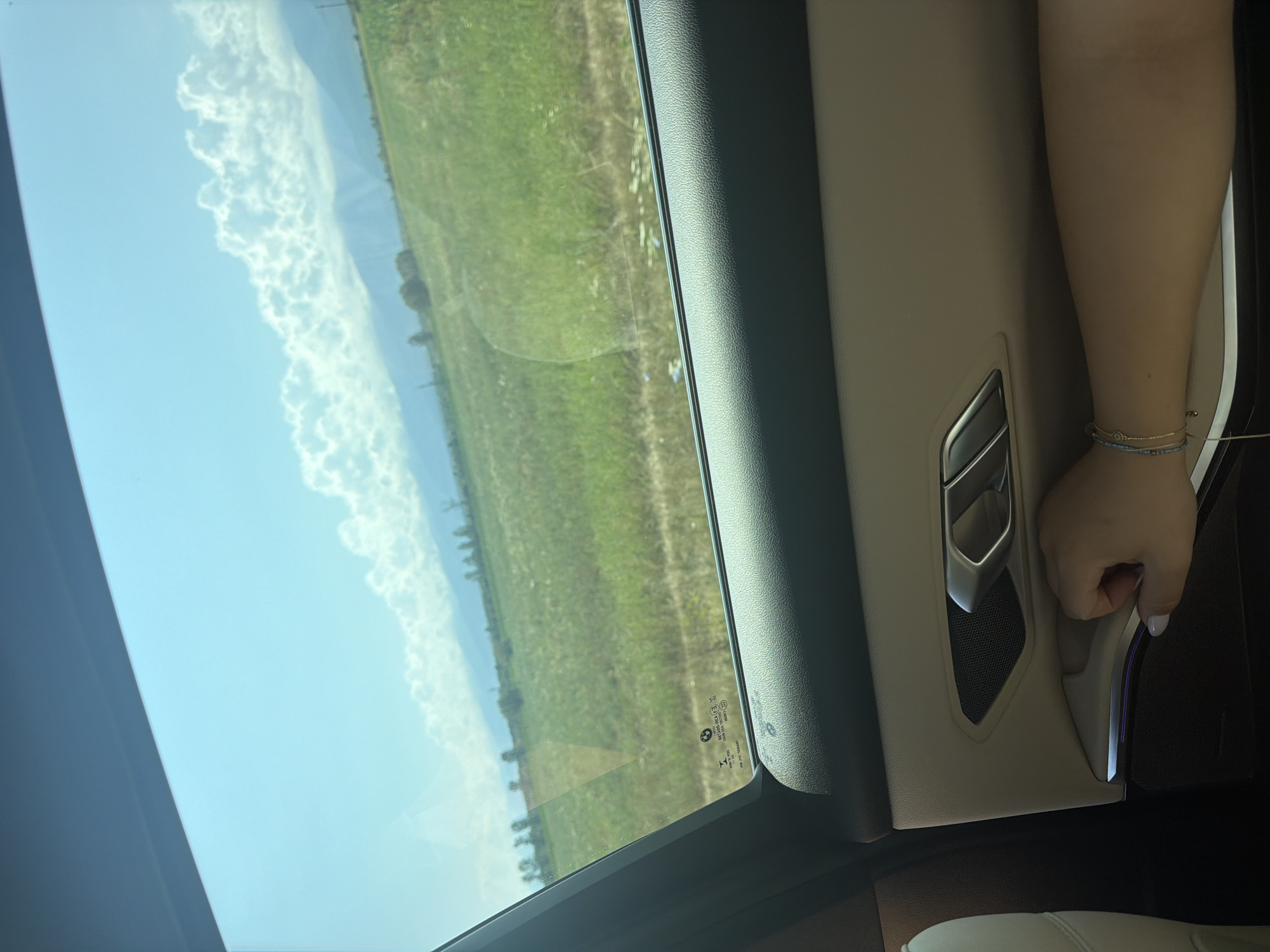 The image, taken from inside a vehicle, shows a view through the window of a rural landscape in Arpașu de Jos, Romania. The foreground features the interior of the car, including the door panel with a handle and a speaker grille. A person's arm and hand, adorned with bracelets, are resting on the door panel. Beyond the window, a vast green field stretches towards rolling hills and distant mountains under a bright blue sky dotted with white clouds. Trees are scattered across the landscape, and a faint reflection is visible on the car window.