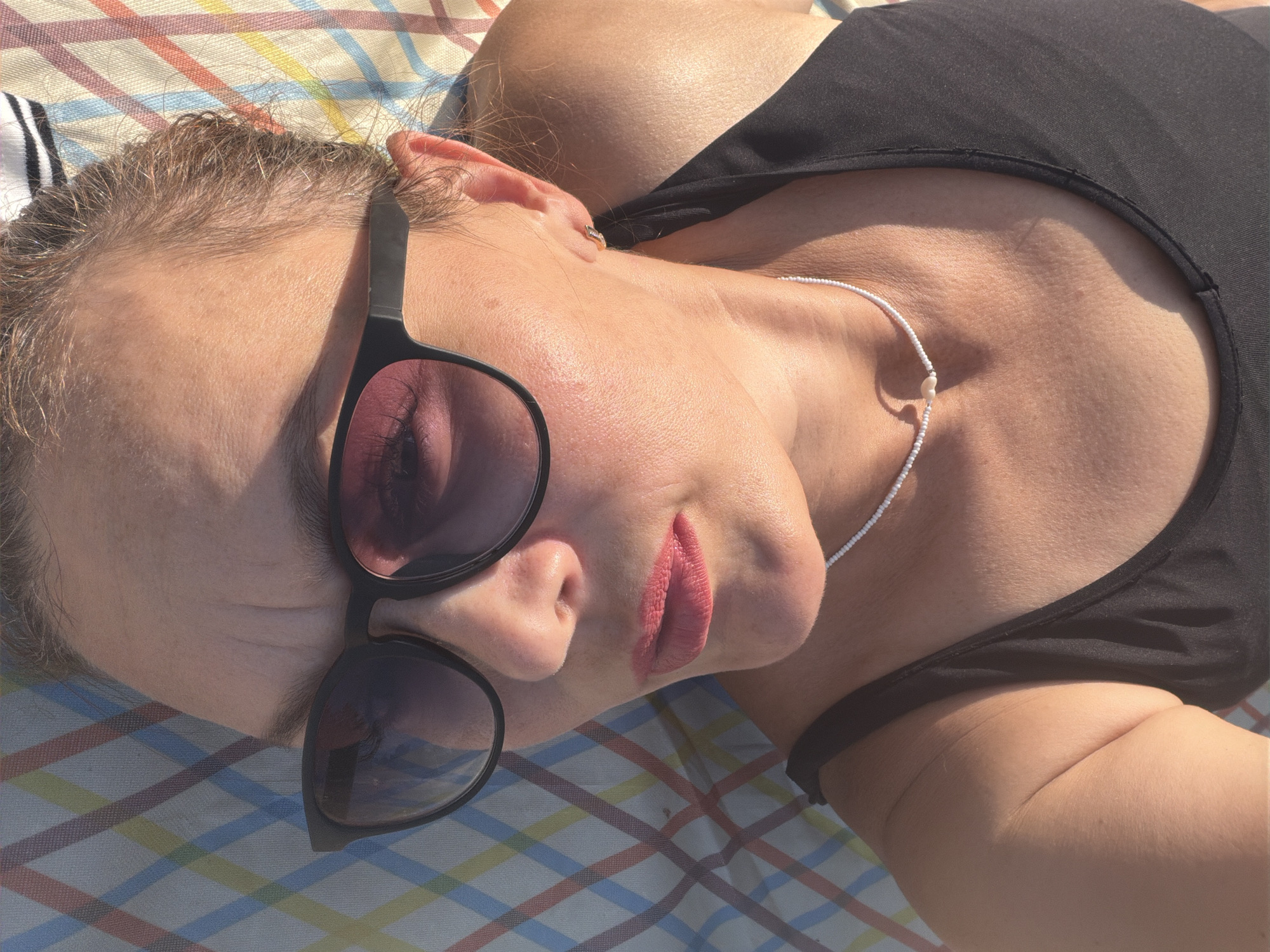 The image shows a close-up, upside-down, angled shot of a person lying on a multi-colored, checkered surface. The person is wearing sunglasses and a black top. Their lips are painted with pink lipstick. A thin, white beaded necklace with a small heart pendant is visible around their neck. The lighting suggests it is daytime, possibly outdoors, with sunlight casting shadows. There are no discernible elements in the image that indicate a specific location within Noviy, Russia.
