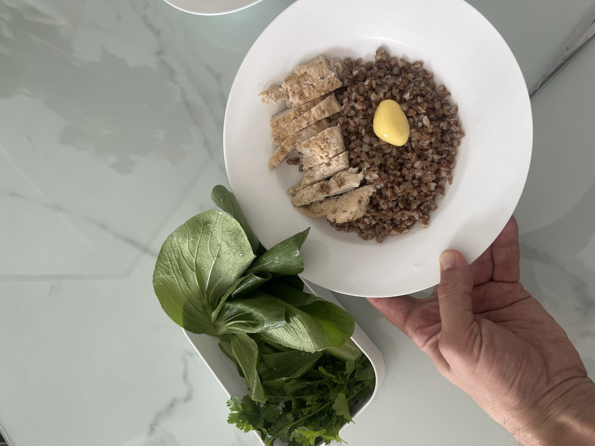 A person's hand holds a white plate containing cooked buckwheat, sliced chicken, and a pat of yellow butter. To the left, a white container holds fresh green leafy vegetables, possibly Bok choy and cilantro. The scene takes place on a white countertop with a marble-like pattern.