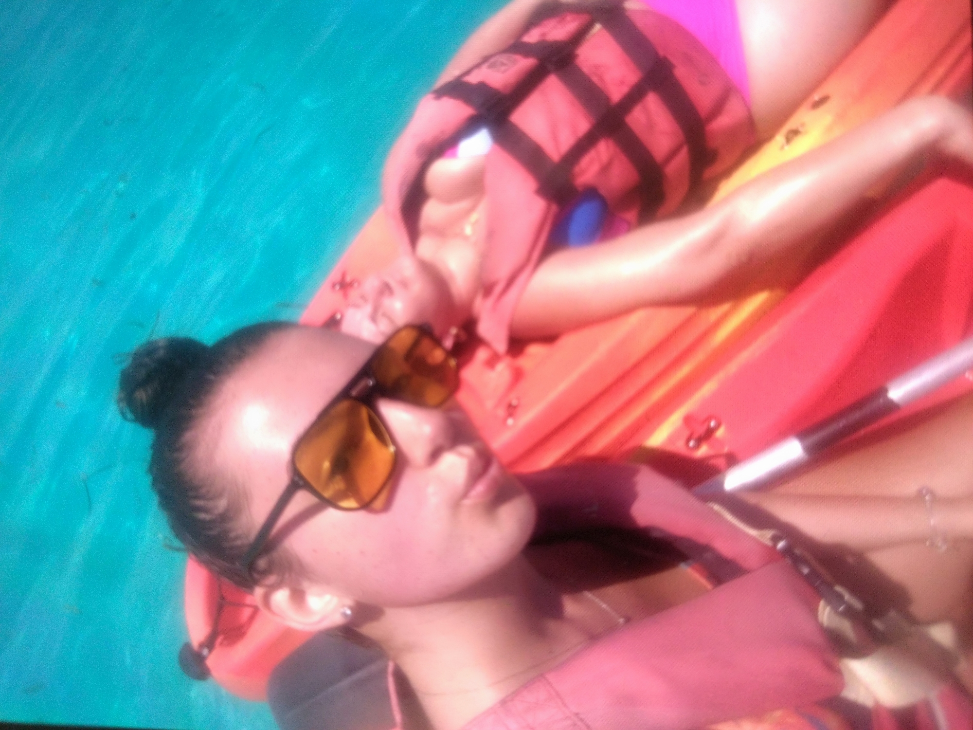 Two people are on a kayak in turquoise water. The person in the foreground is wearing sunglasses and a life jacket. The person in the background is also wearing a life jacket. A paddle is visible. The location is not discernable from the image.